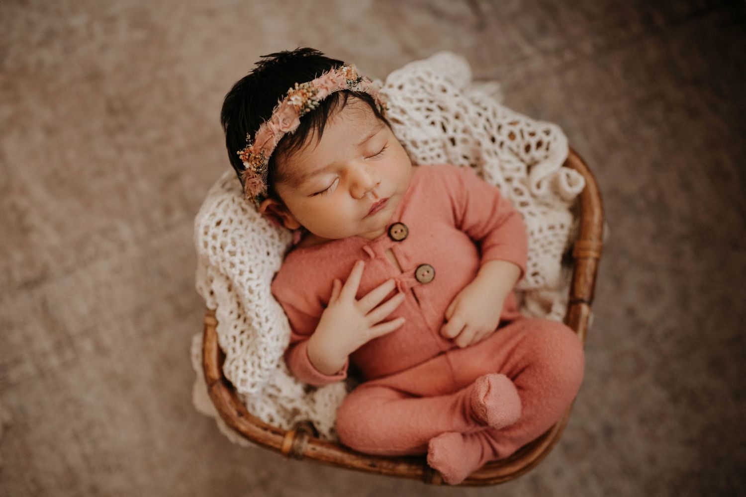 Sleeping newborn wearing coral pink outfit rests peacefully in wicker basket with lace blanket and floral headband.