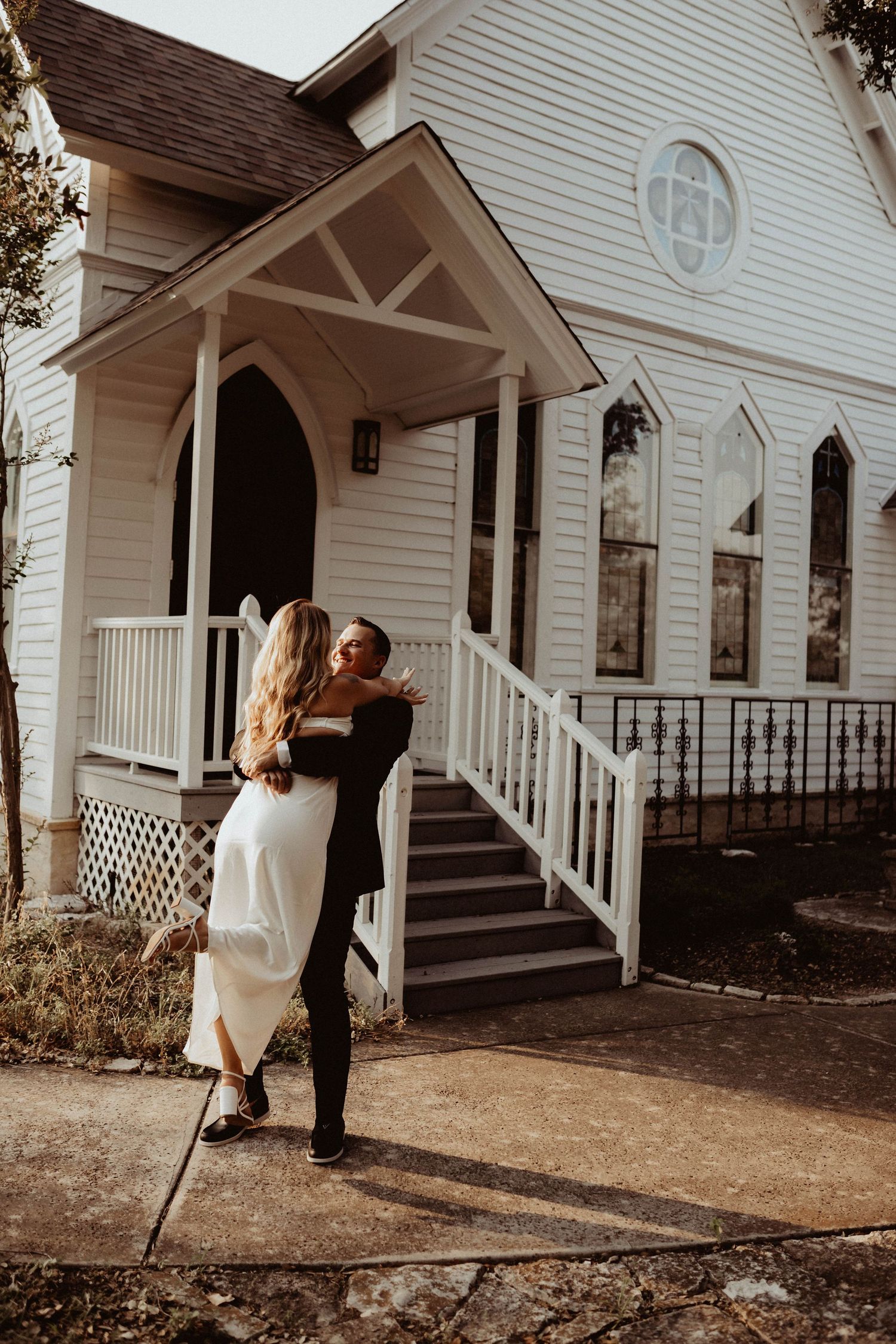 A couple shares a romantic moment outside a charming white chapel with a rustic porch and steps.