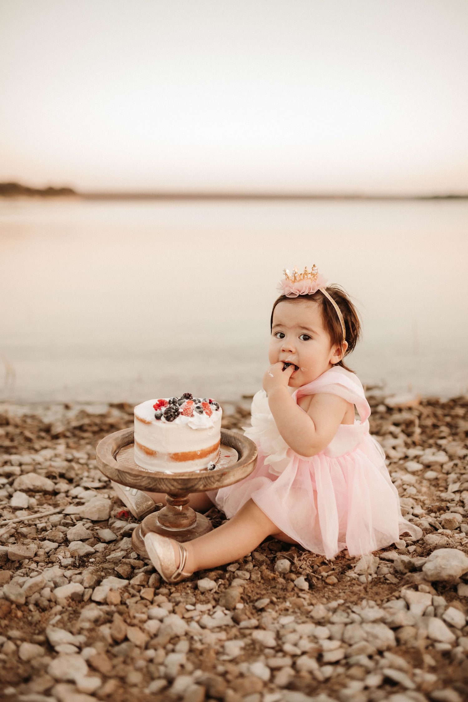 A small child in a pink tutu dress sits on a rocky beach near a decorated birthday cake.