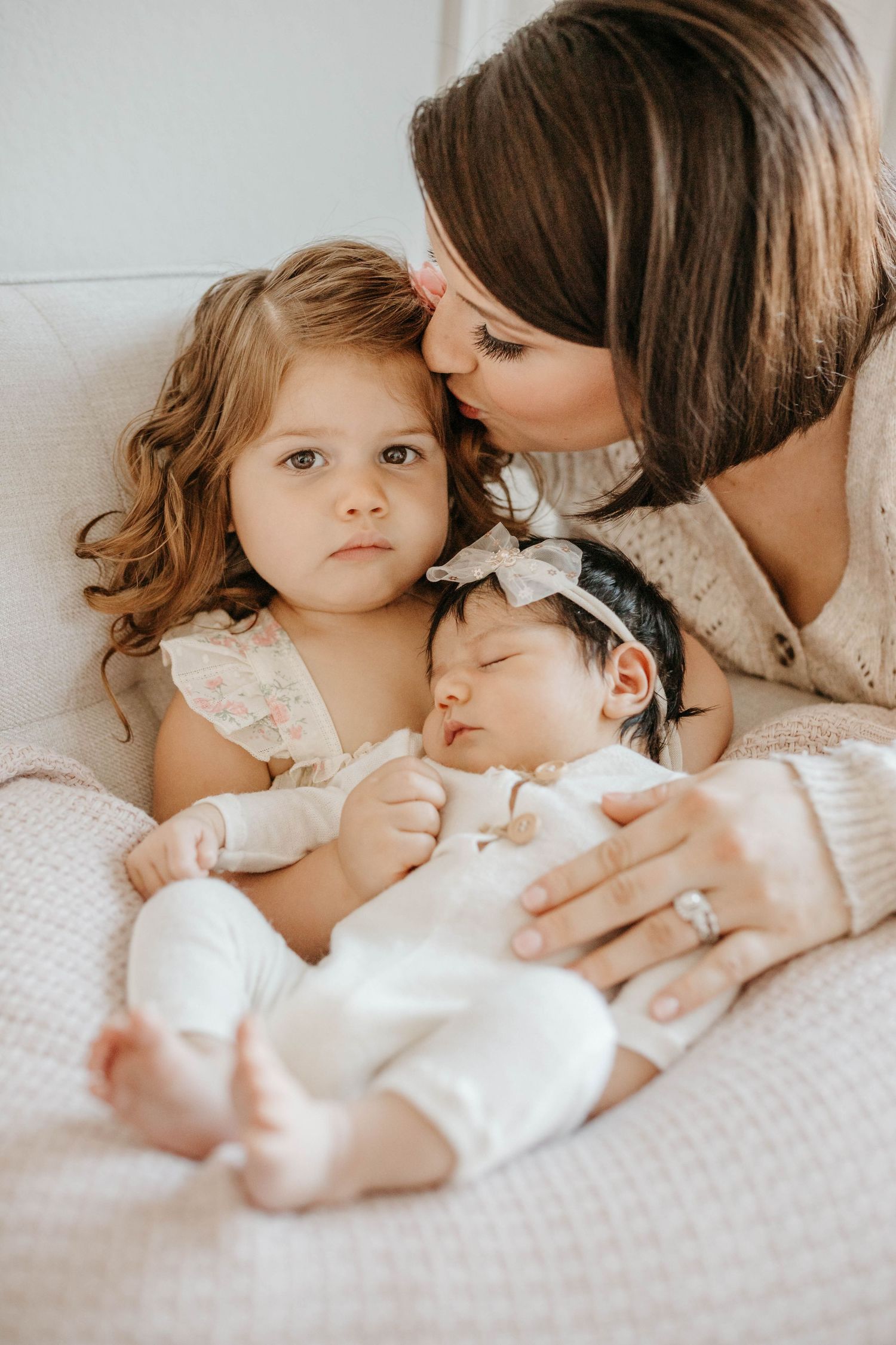 Tender family moment captured in soft beige tones as siblings share affectionate cuddles on white bedding.