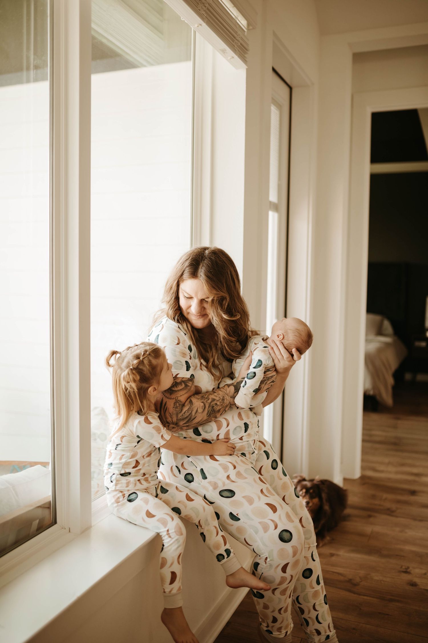 A mother in a floral dress dances with her baby by a sunlit window in a minimalist room with wooden floors.