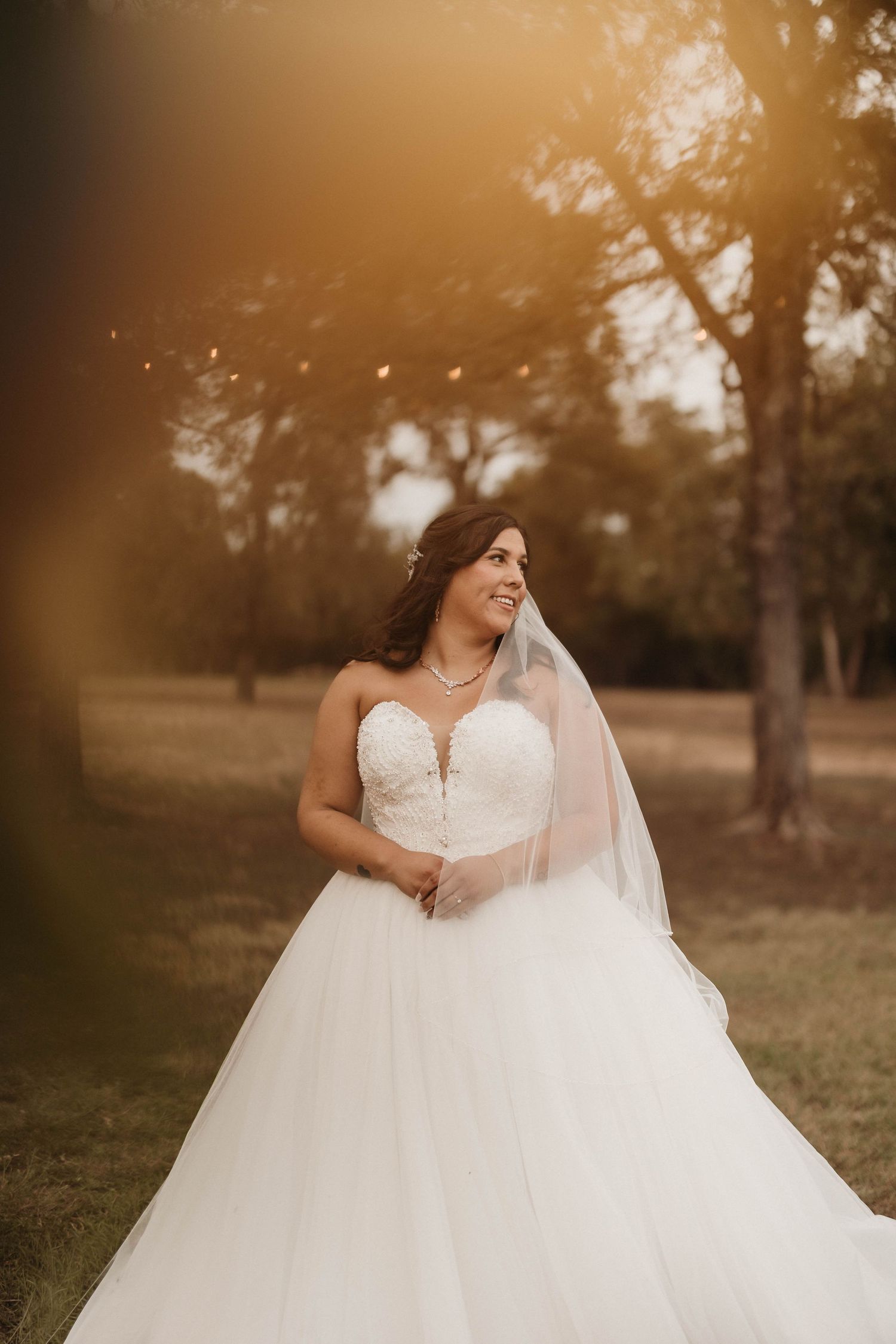 A bride in a flowing white ballgown twirls joyfully in a sunlit outdoor setting at golden hour.