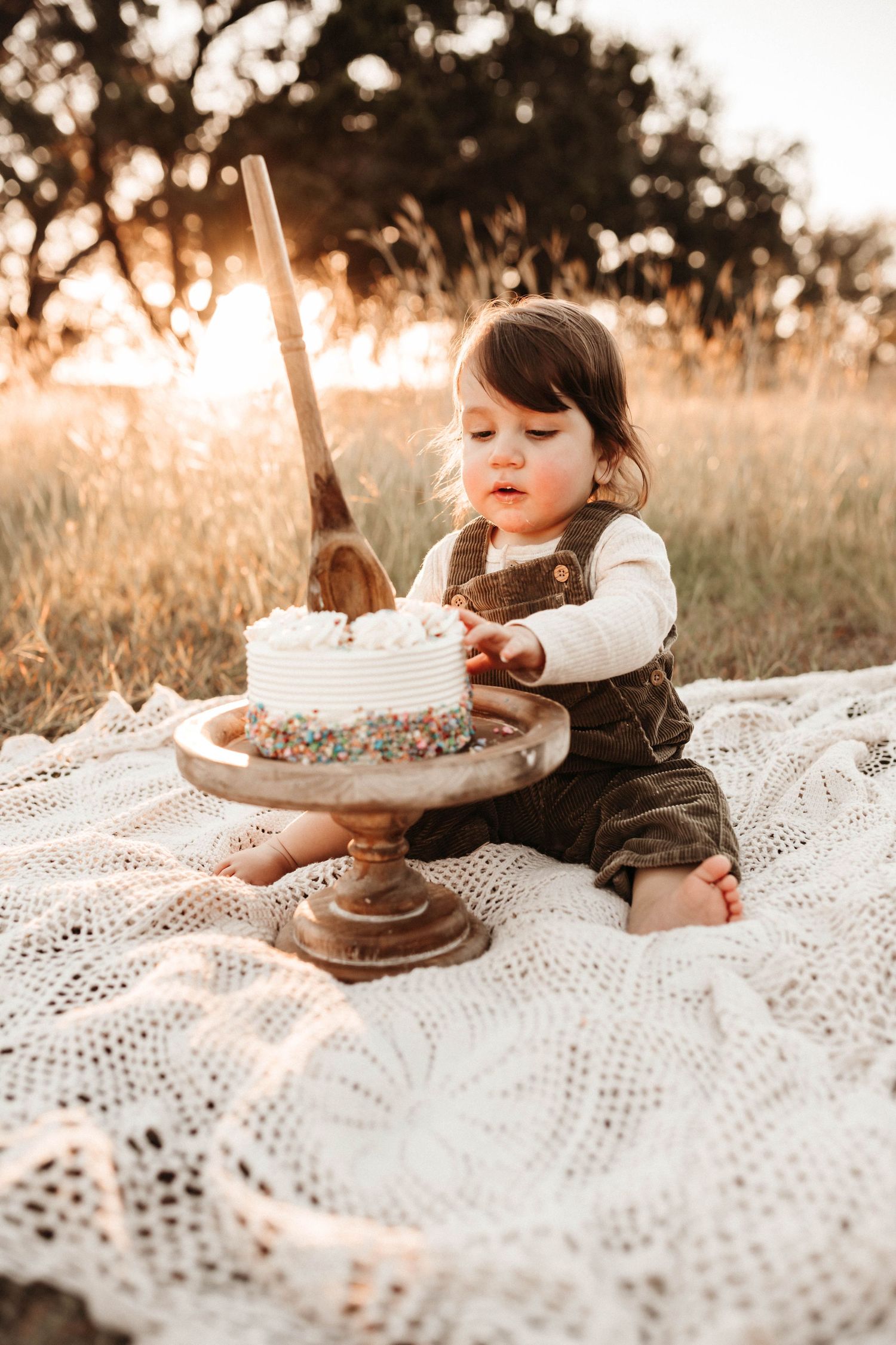 A small child in overalls sits on a lace blanket reaching for a cake on a pedestal during golden hour sunset.