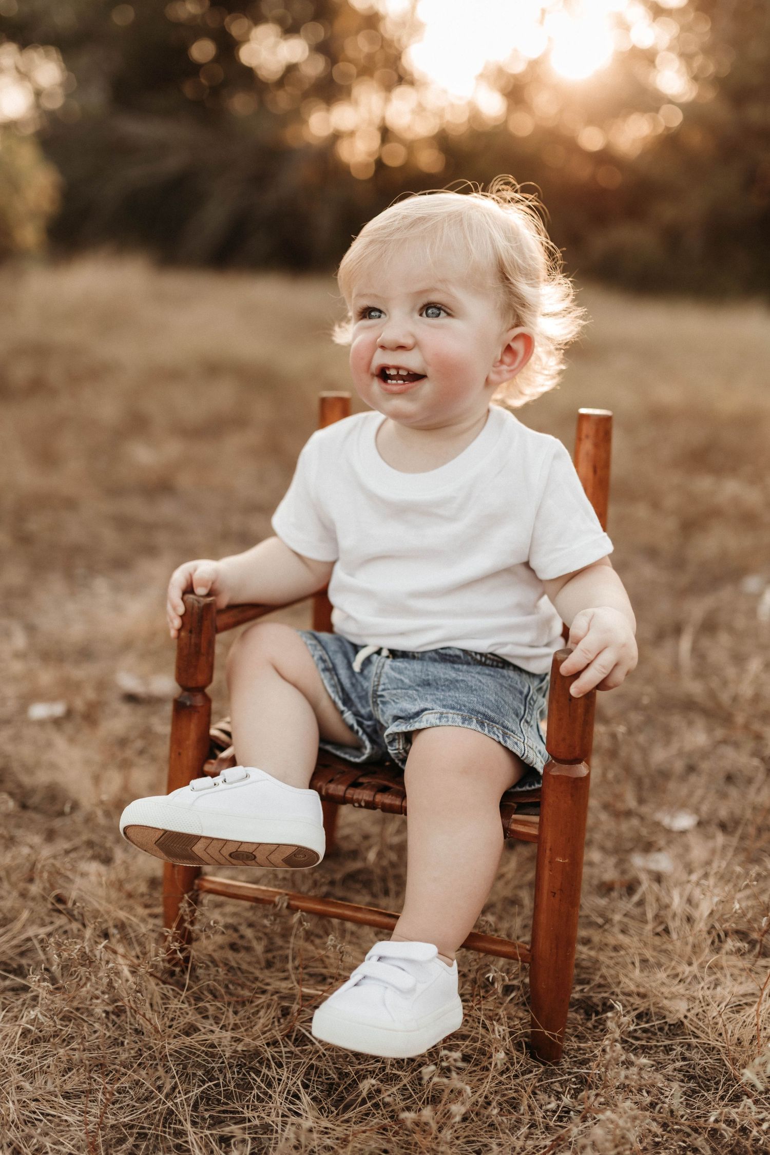 A baby in a white shirt and denim shorts sits on a small wooden chair in a sunlit field at golden hour.