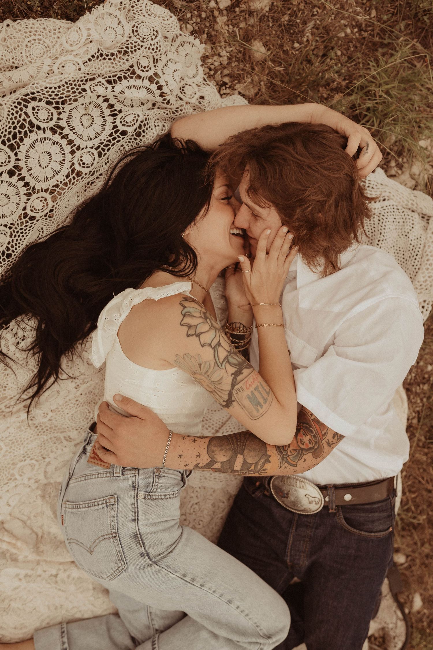 A couple sharing an intimate moment on lace fabric with warm sepia tones and visible tattoos.