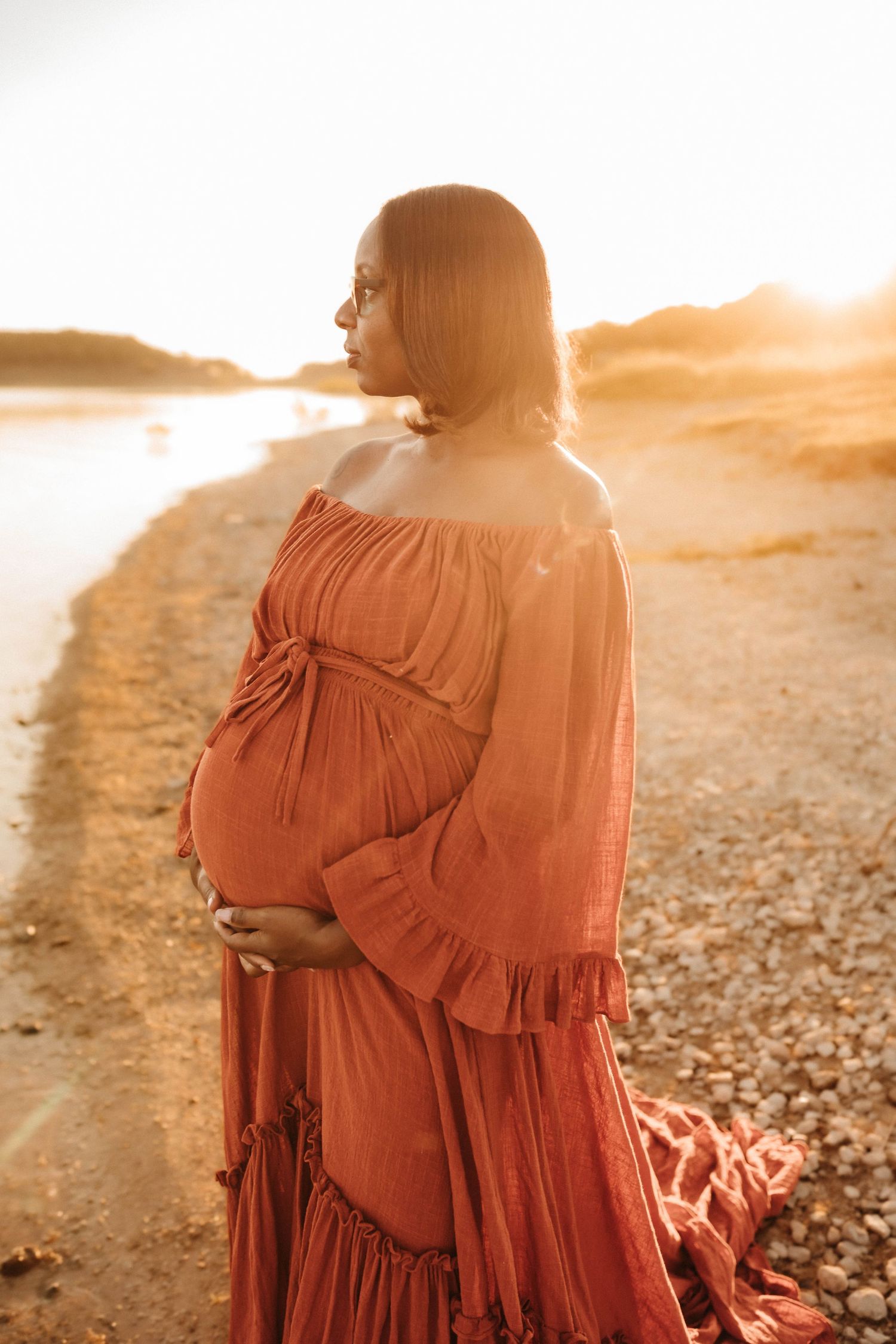 A silhouetted figure in a flowing rust-colored dress stands on a rocky beach at sunset, creating a dreamy atmosphere.
