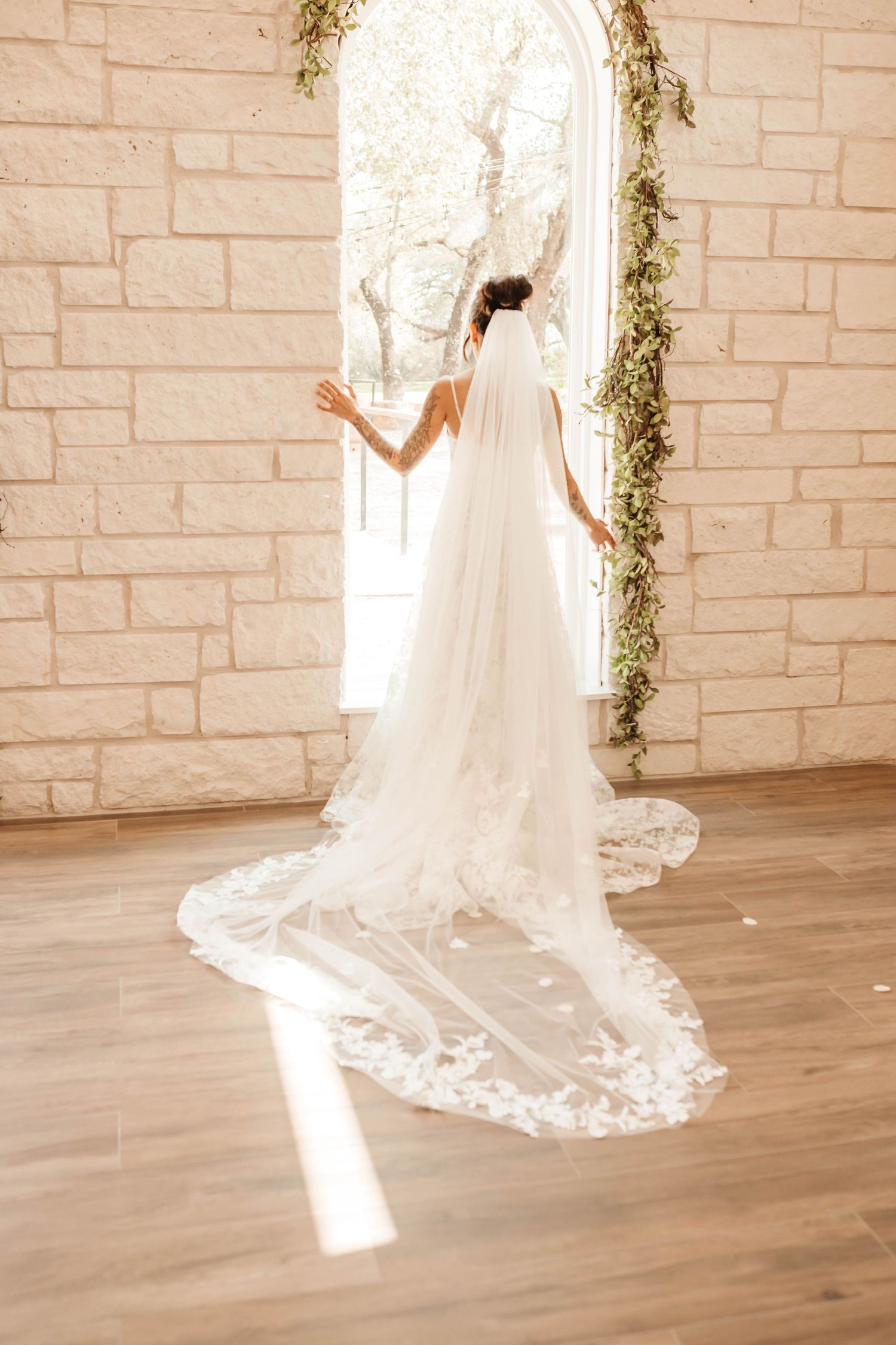 A bride in a flowing white lace veil stands in front of an arched window with natural light streaming in.