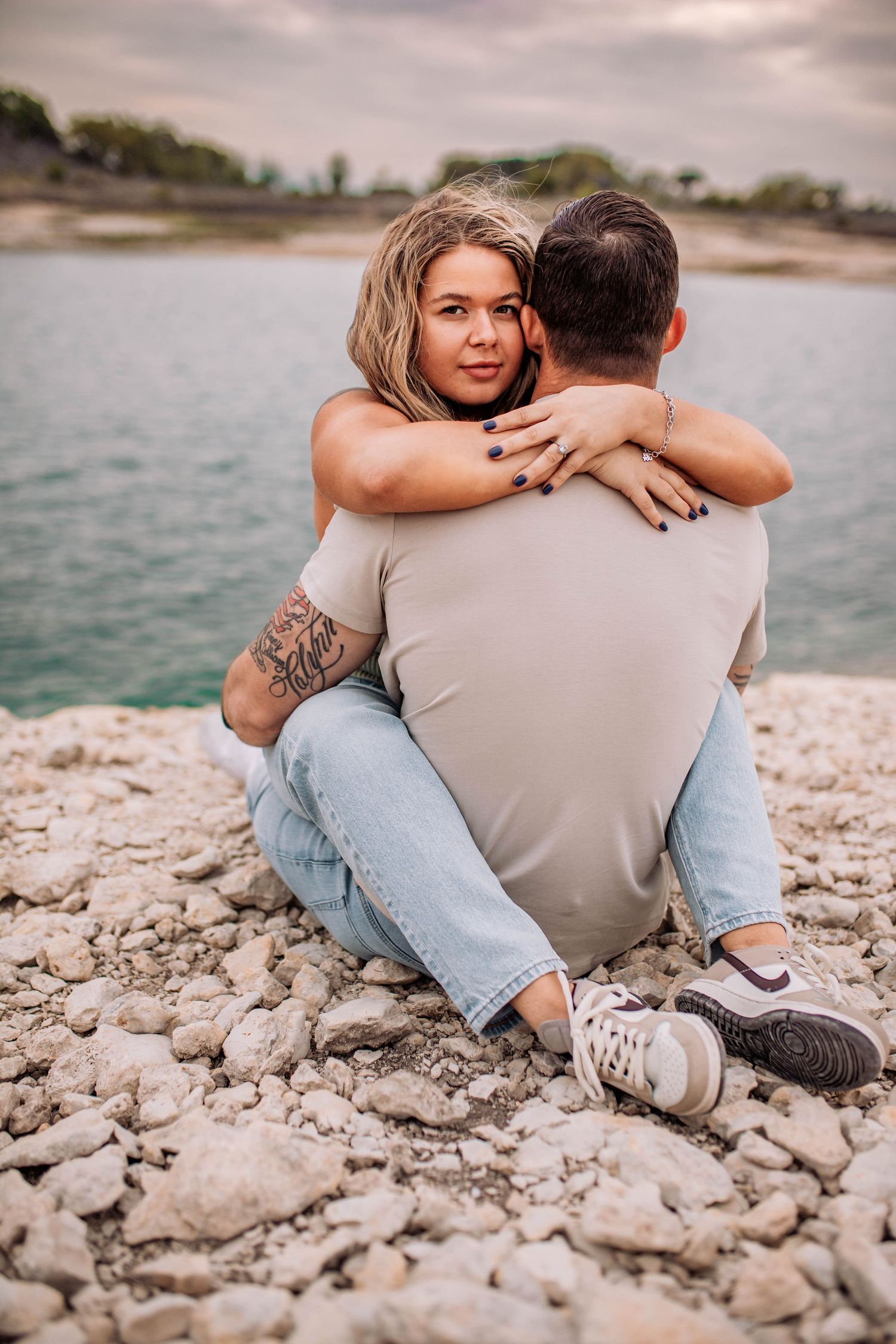 A couple sits together on rocky shoreline overlooking a serene lake at sunset.