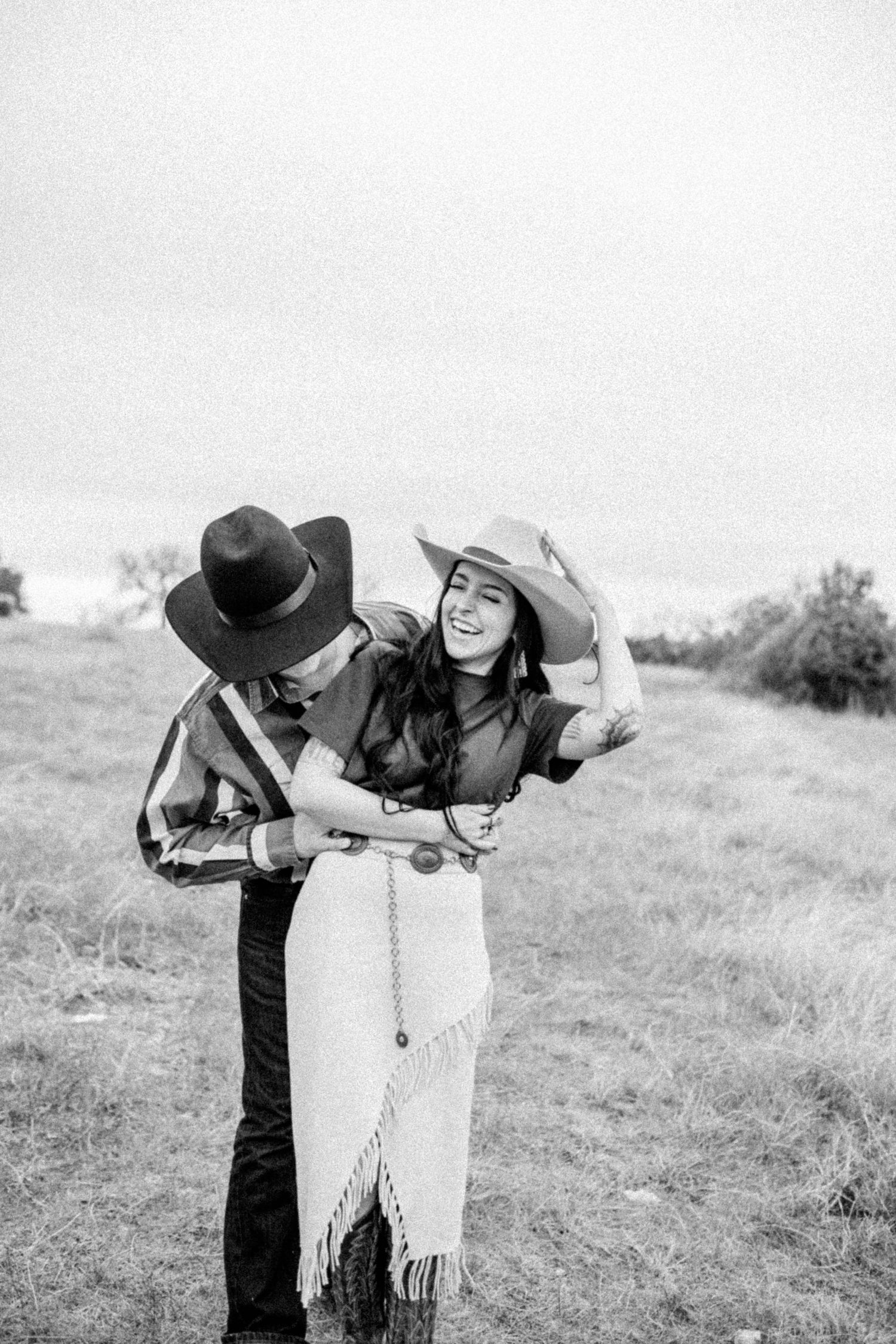 Black and white photo of a playful western-themed couple posing in a grassy field.