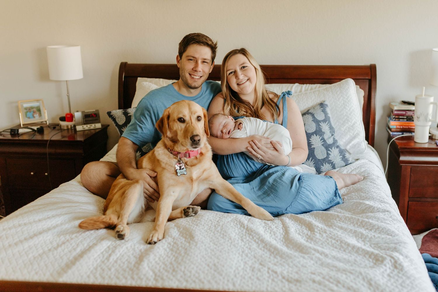 A family relaxes together on a bed with their golden retriever and newborn baby in a cozy bedroom setting.