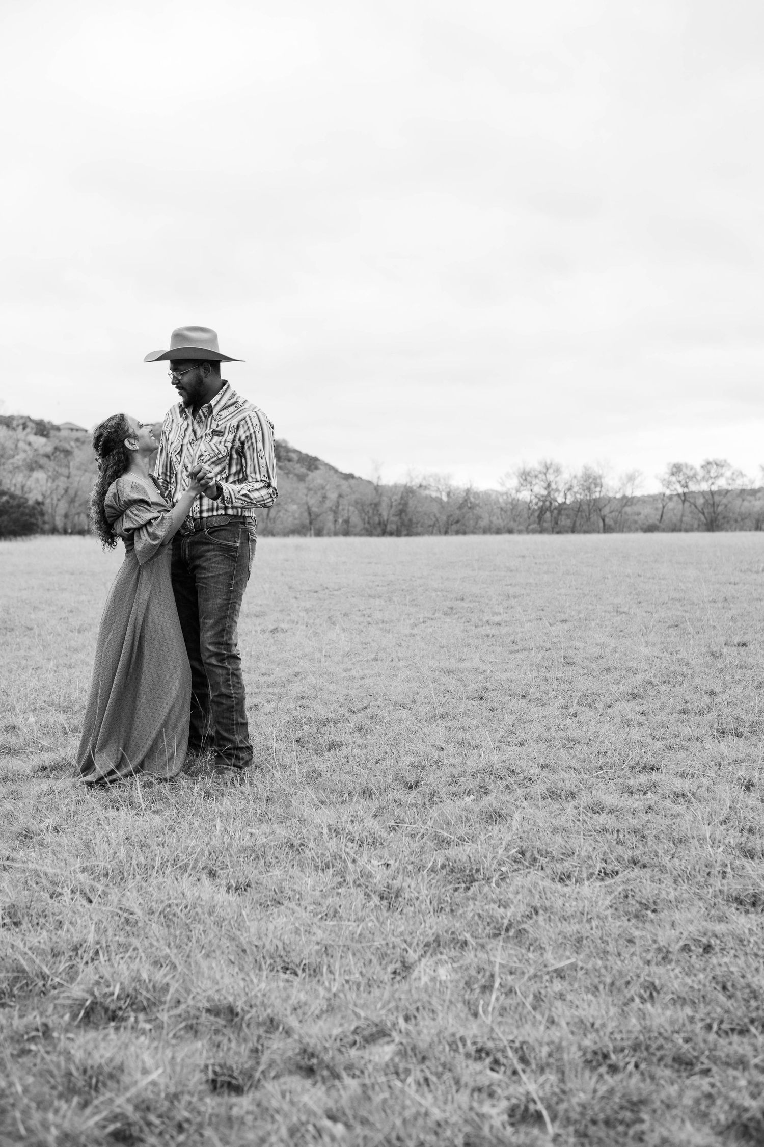 Black and white photo of a couple embracing in a serene open field with mountains in the background.