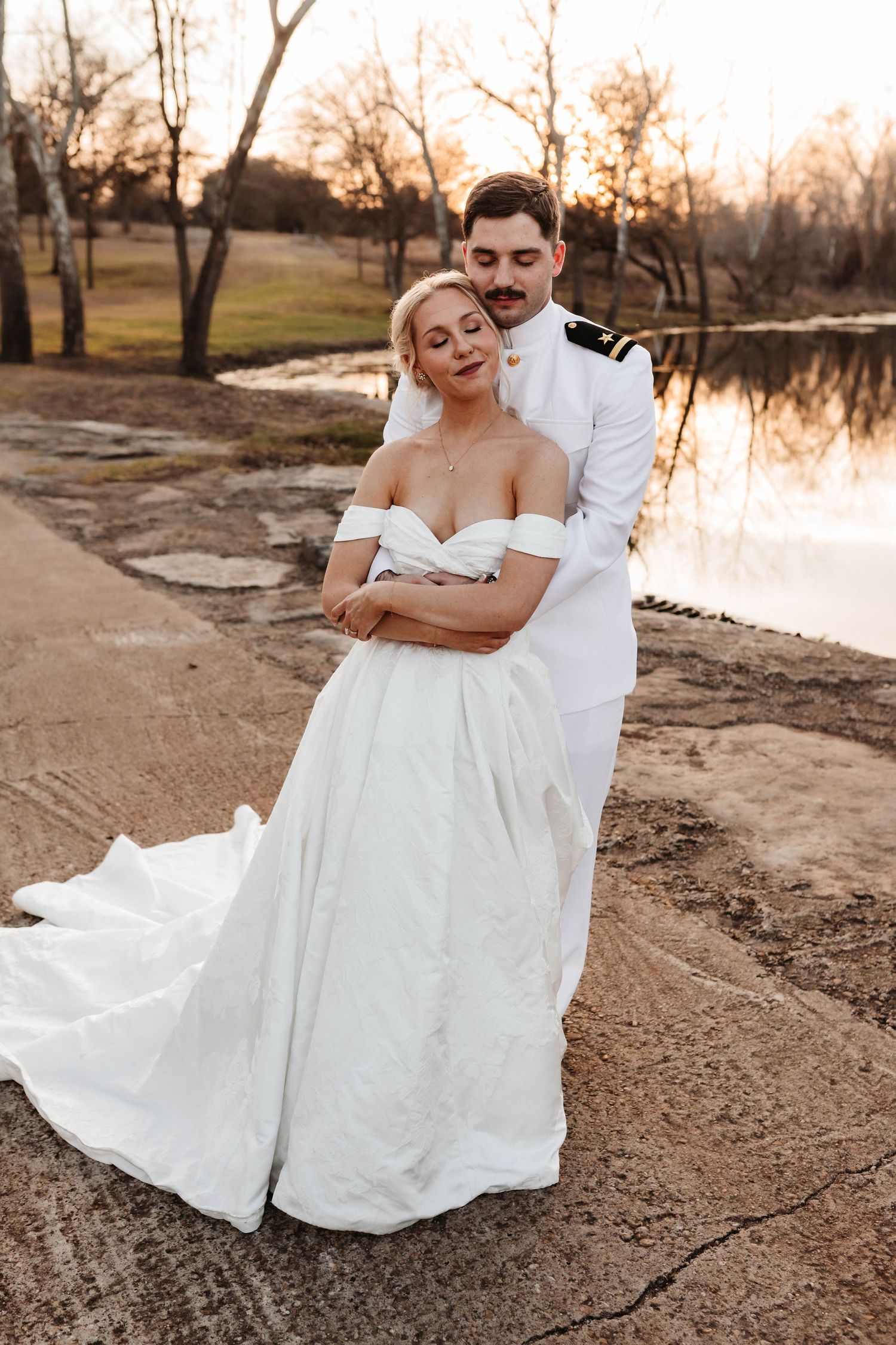 A couple in wedding attire shares a romantic moment by a lake at sunset while sitting on sandy ground.