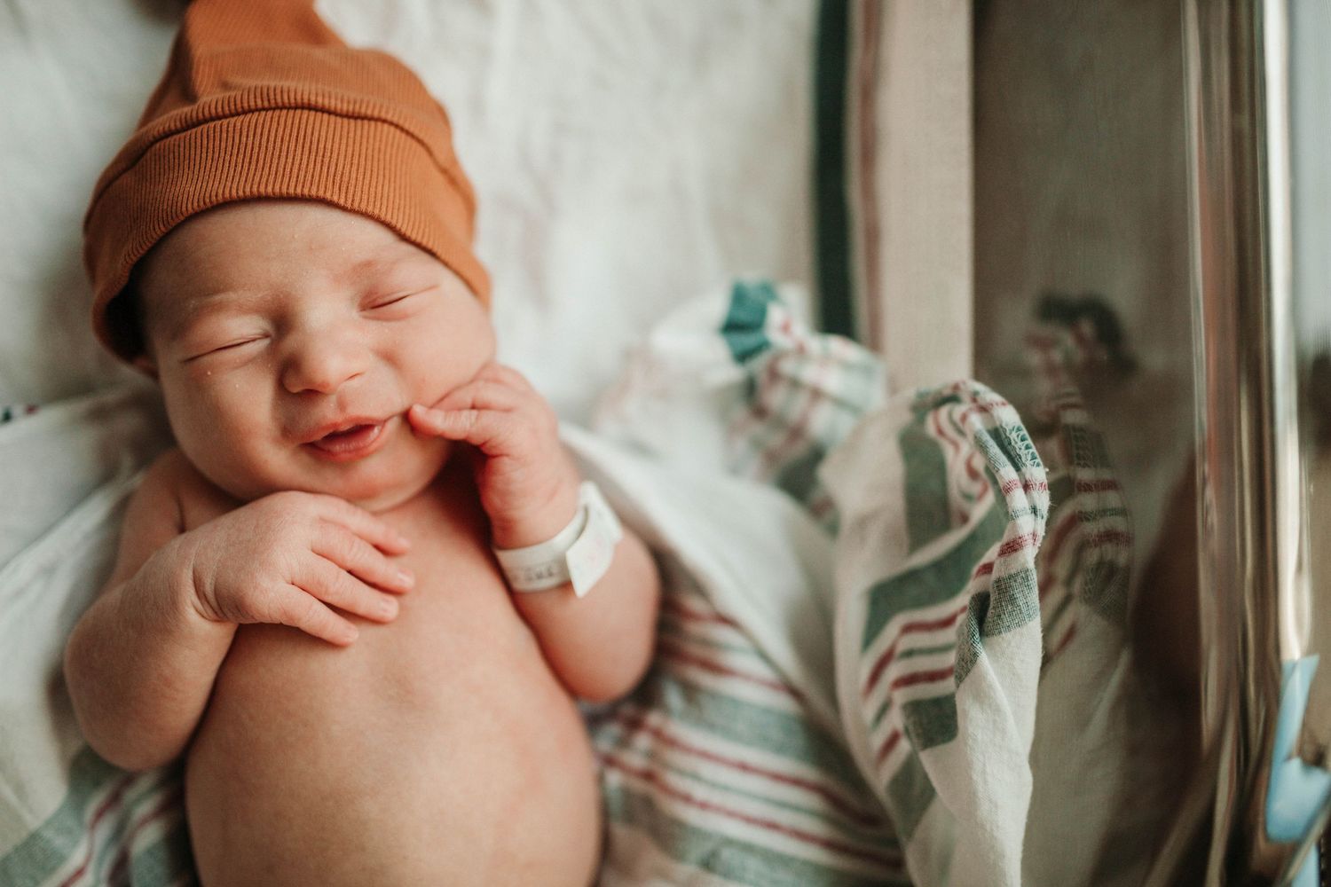 A baby wearing an orange beanie hat lies on a white blanket near a window with natural light.
