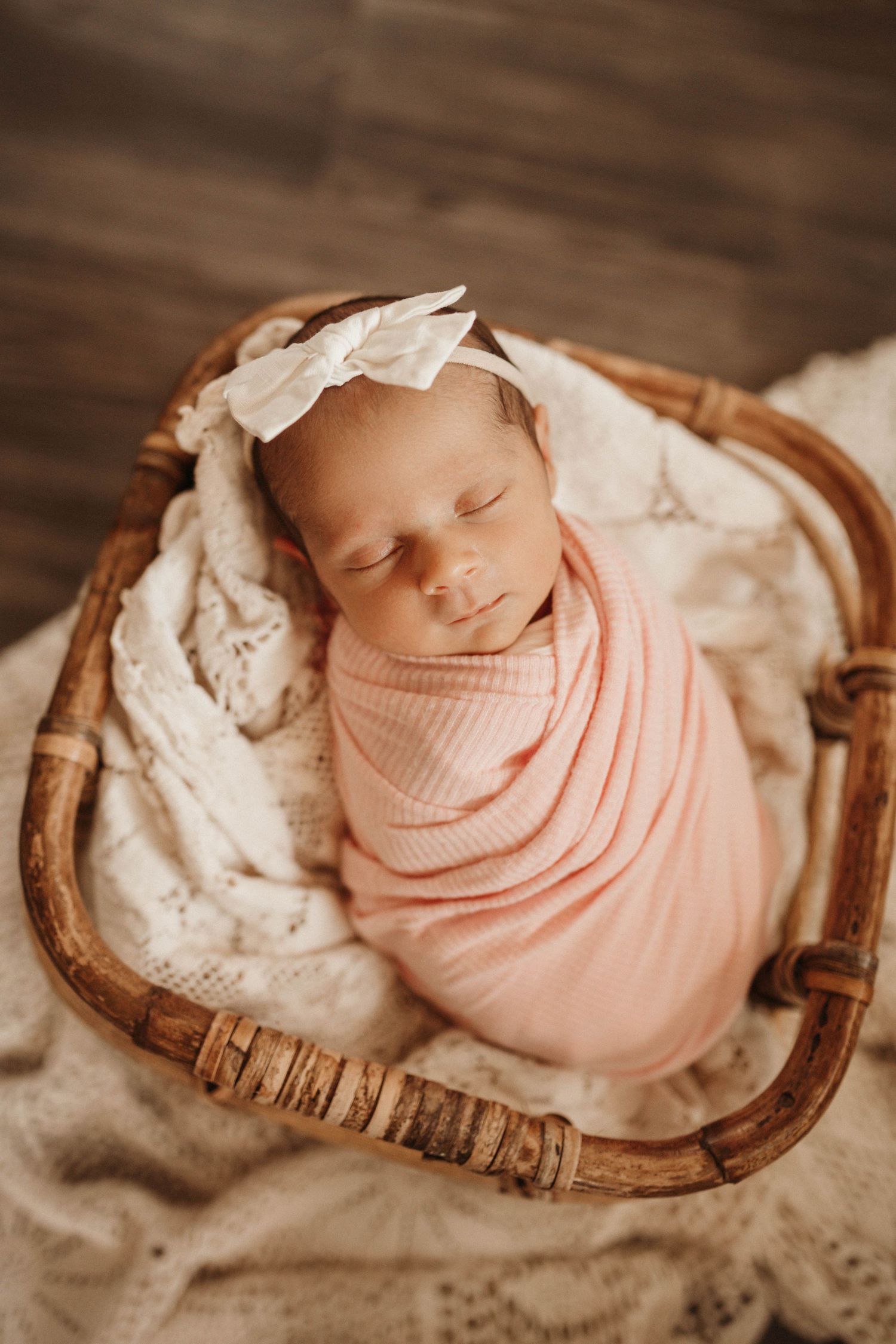 A sleeping newborn wrapped in soft pink swaddle lies peacefully on a rustic wooden surface with delicate lace details.