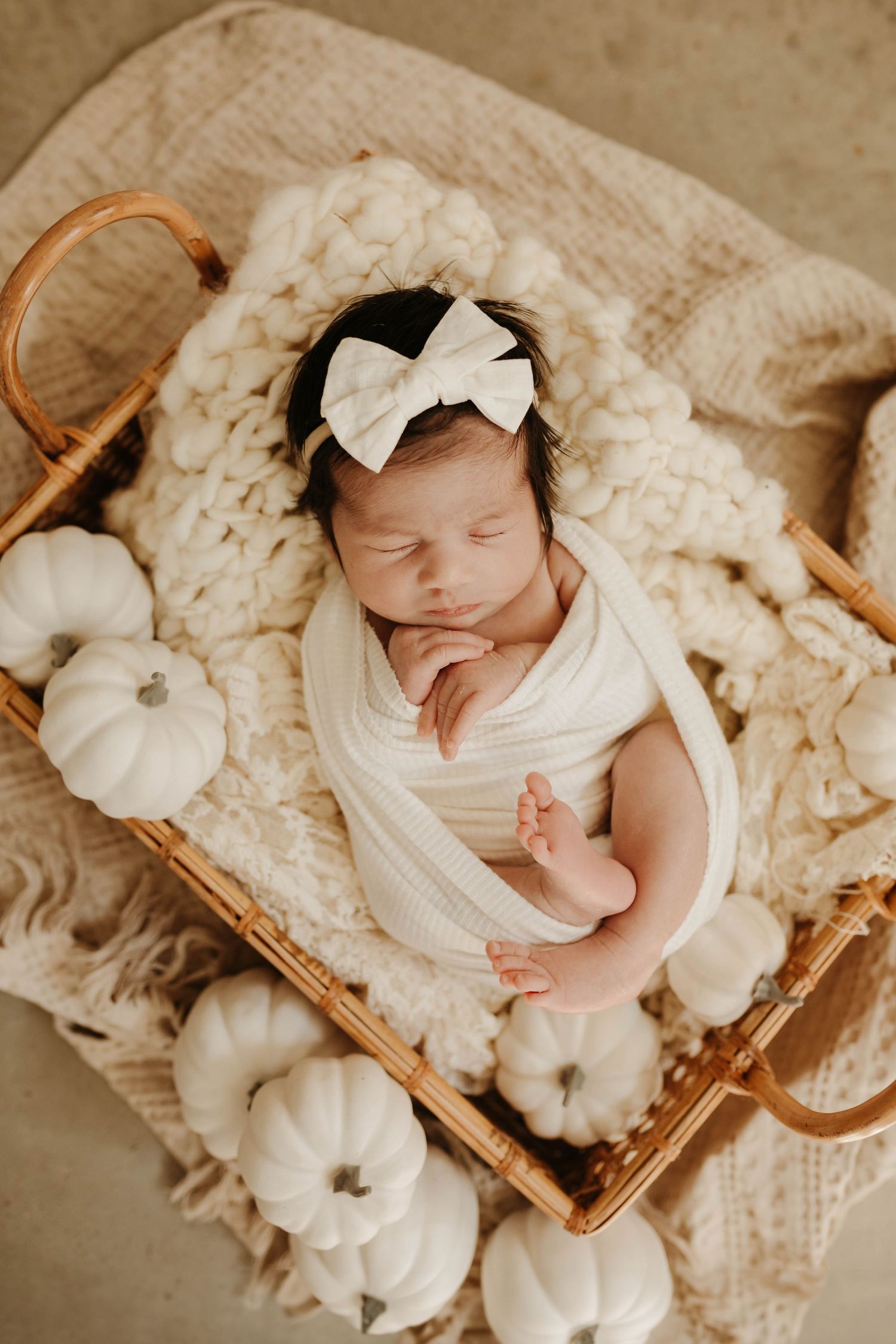 A sleeping newborn in white wrap lies in a woven basket surrounded by small white decorative pumpkins on cream textured fabric.