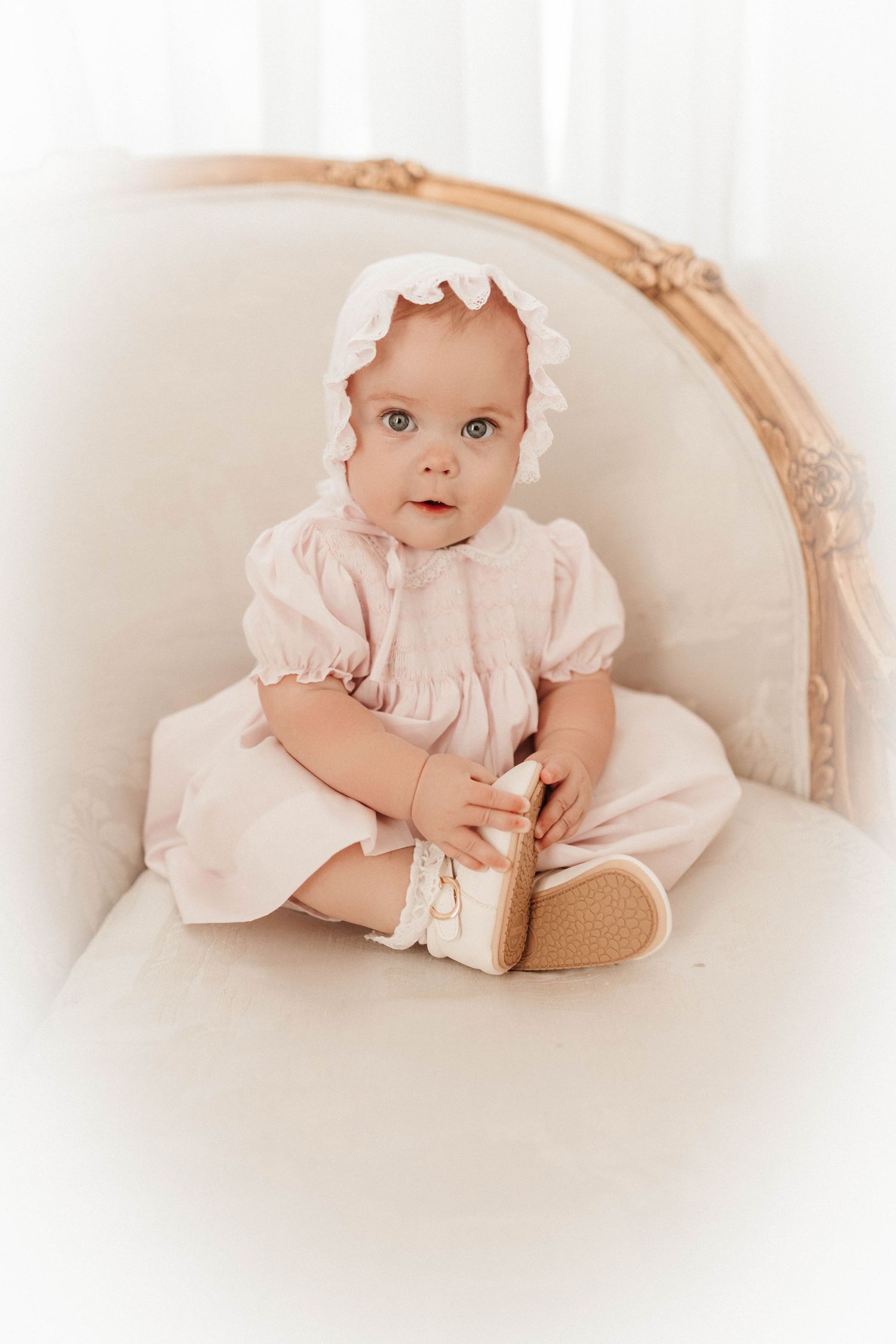 Sweet baby in pink ruffled dress laying on cream colored antique chaise lounge in soft dreamy photography style.