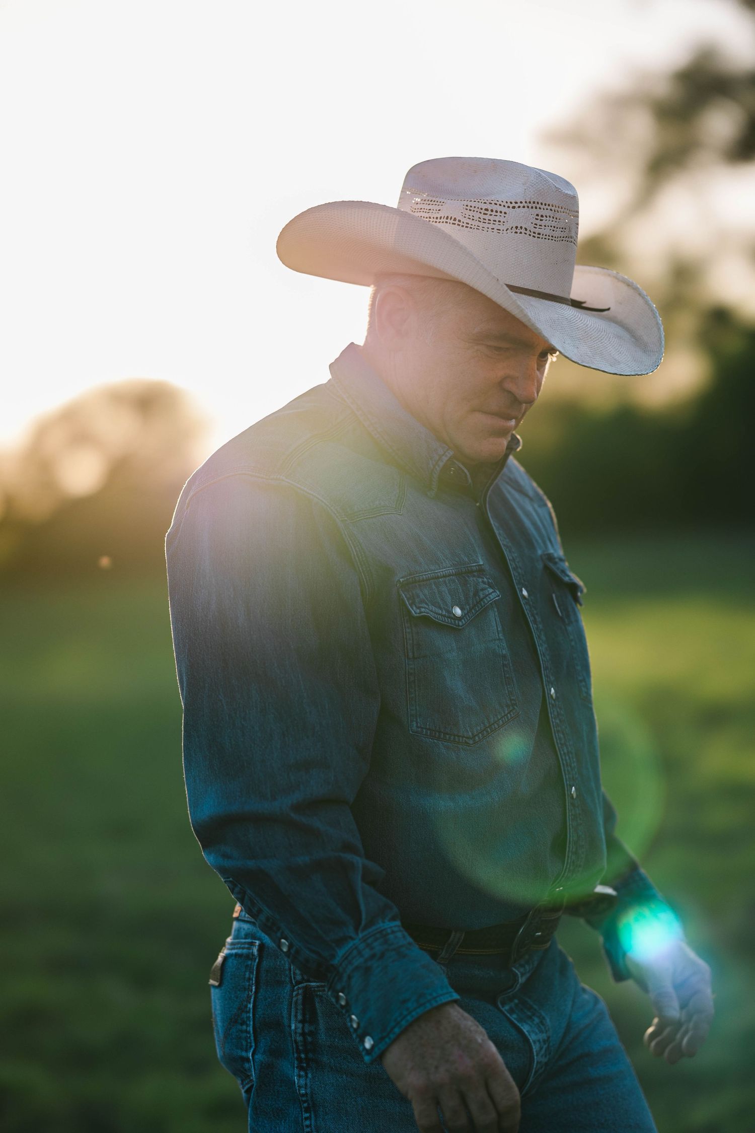 Rugged cowboy in a white hat walking through a sunlit green field with lens flare at sunset.