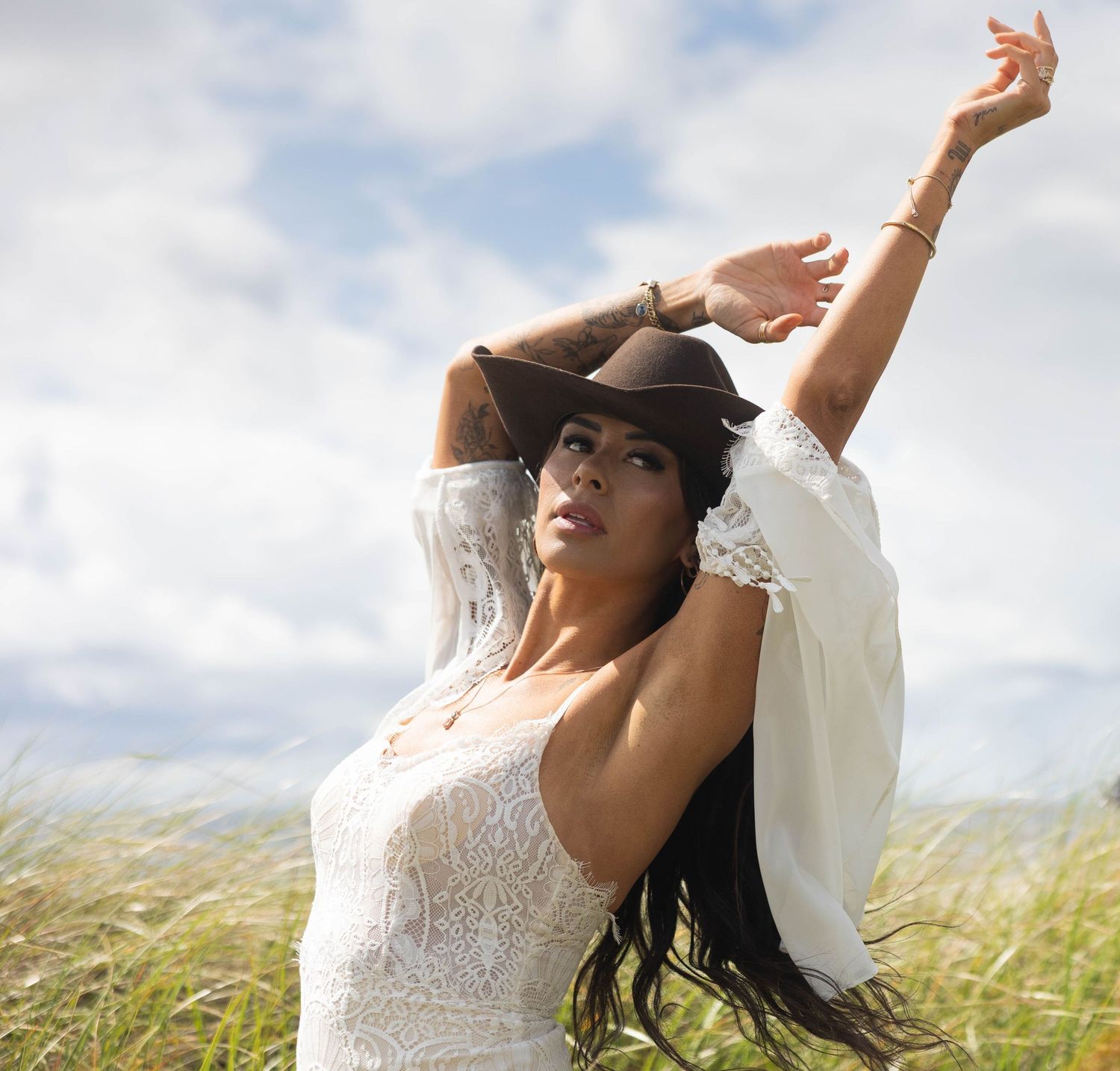 Confident woman in white lace dress and cowboy hat posing in sunny meadow with arms raised.