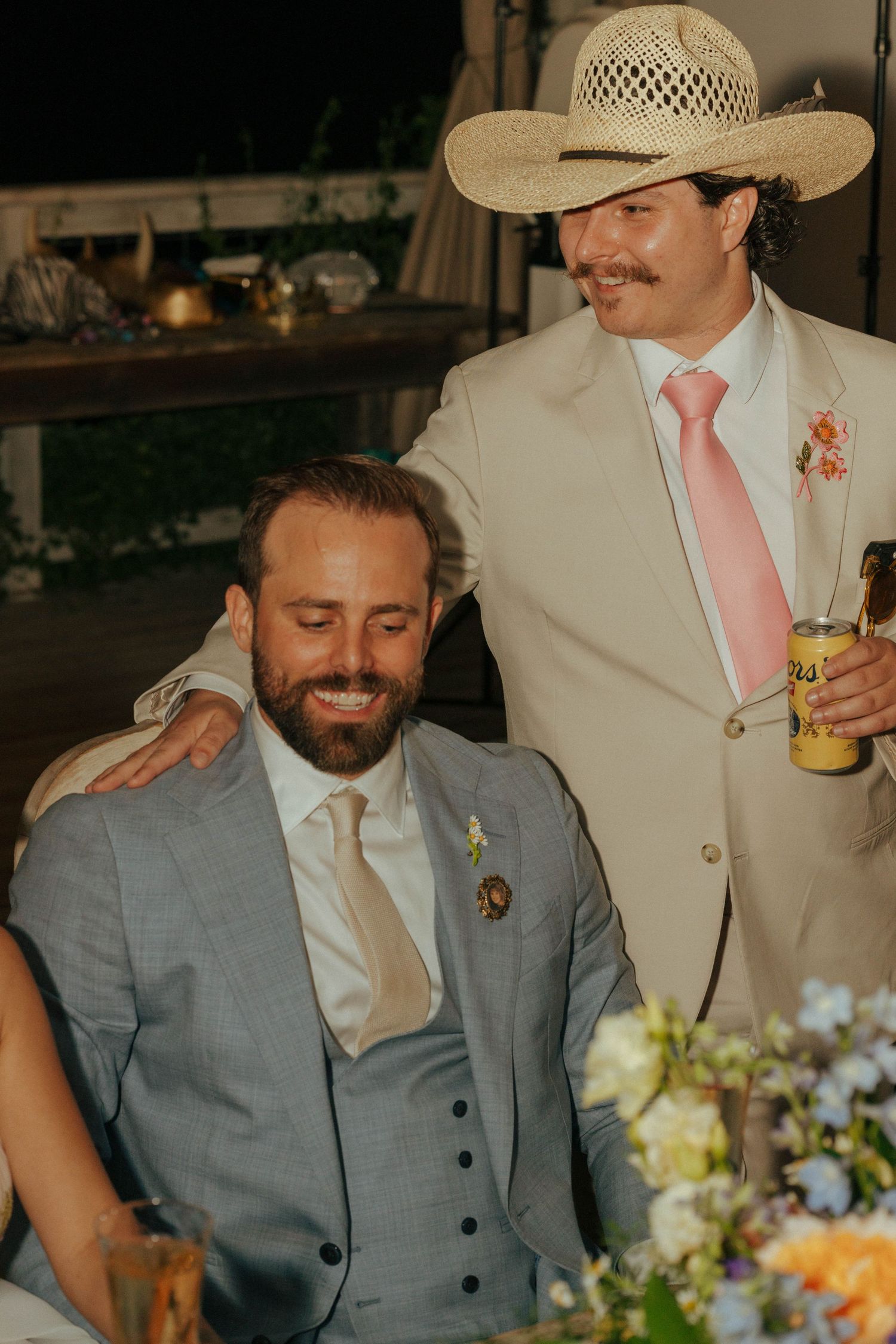 Man in cowboy hat and pink tie places hand on seated grooms shoulder during outdoor wedding reception toast.