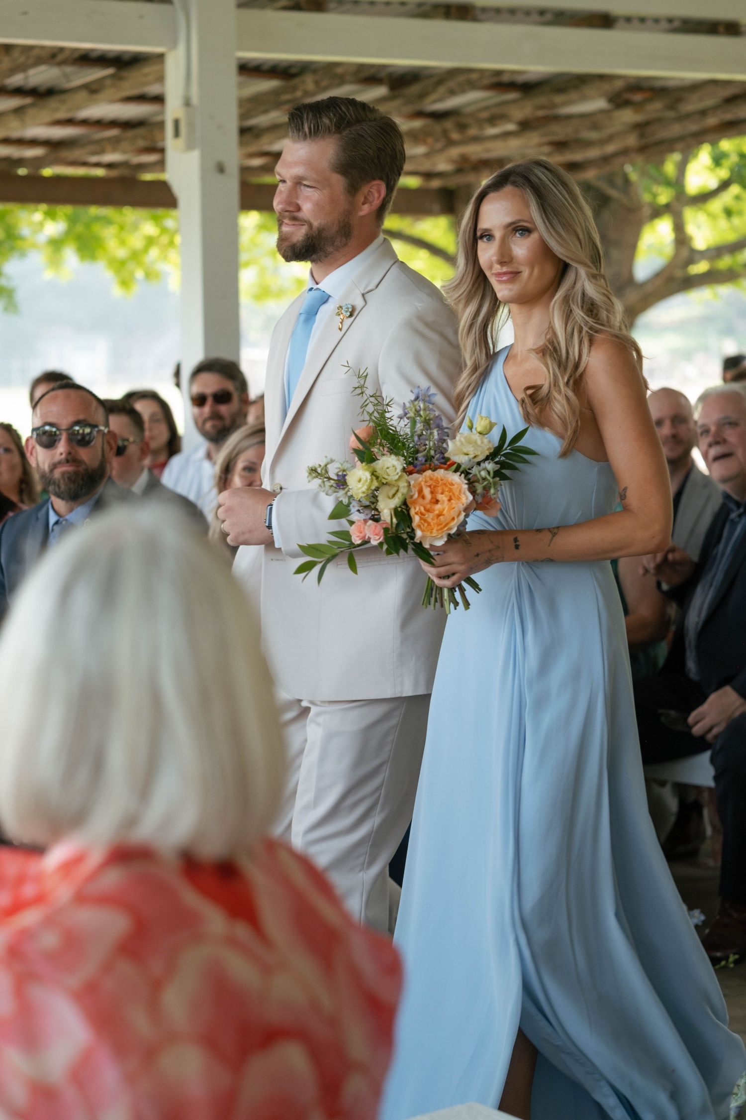 Groomsman in beige suit walks bridesmaid in light blue gown down ceremony aisle.
