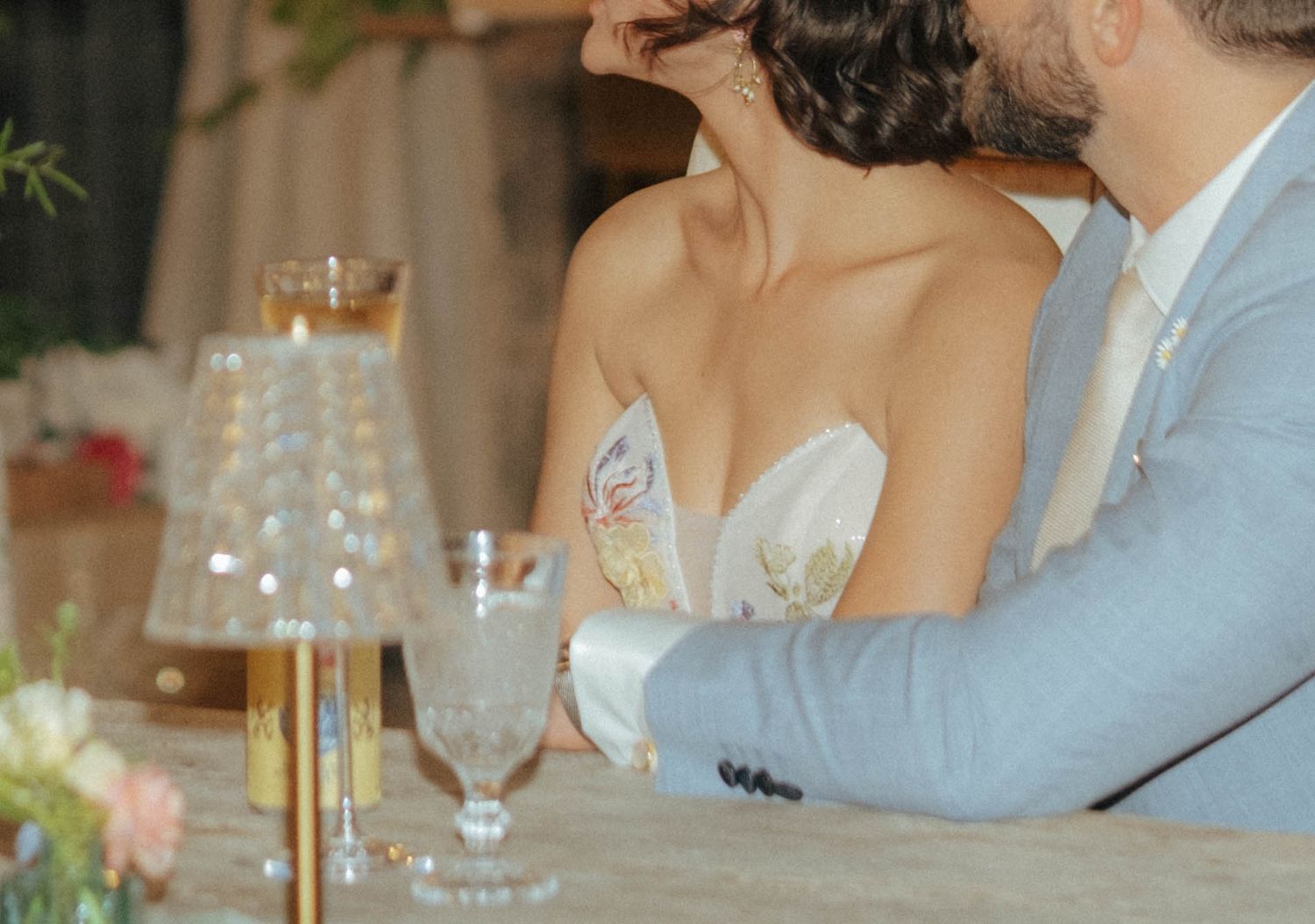 Bride in floral strapless gown leans on groom in grey suit at elegantly decorated wedding reception table.