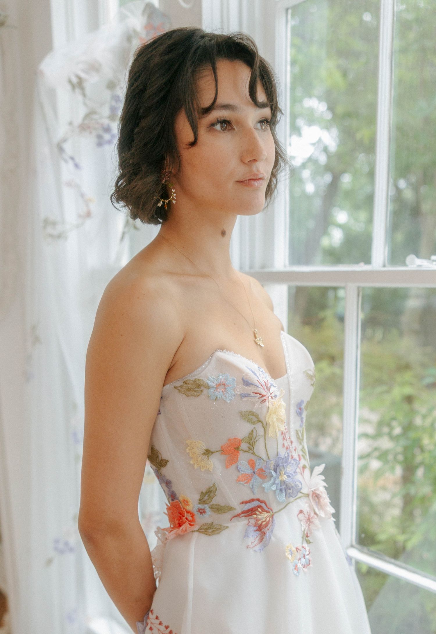 Bride in a strapless floral embroidered wedding dress standing by a window in soft light.