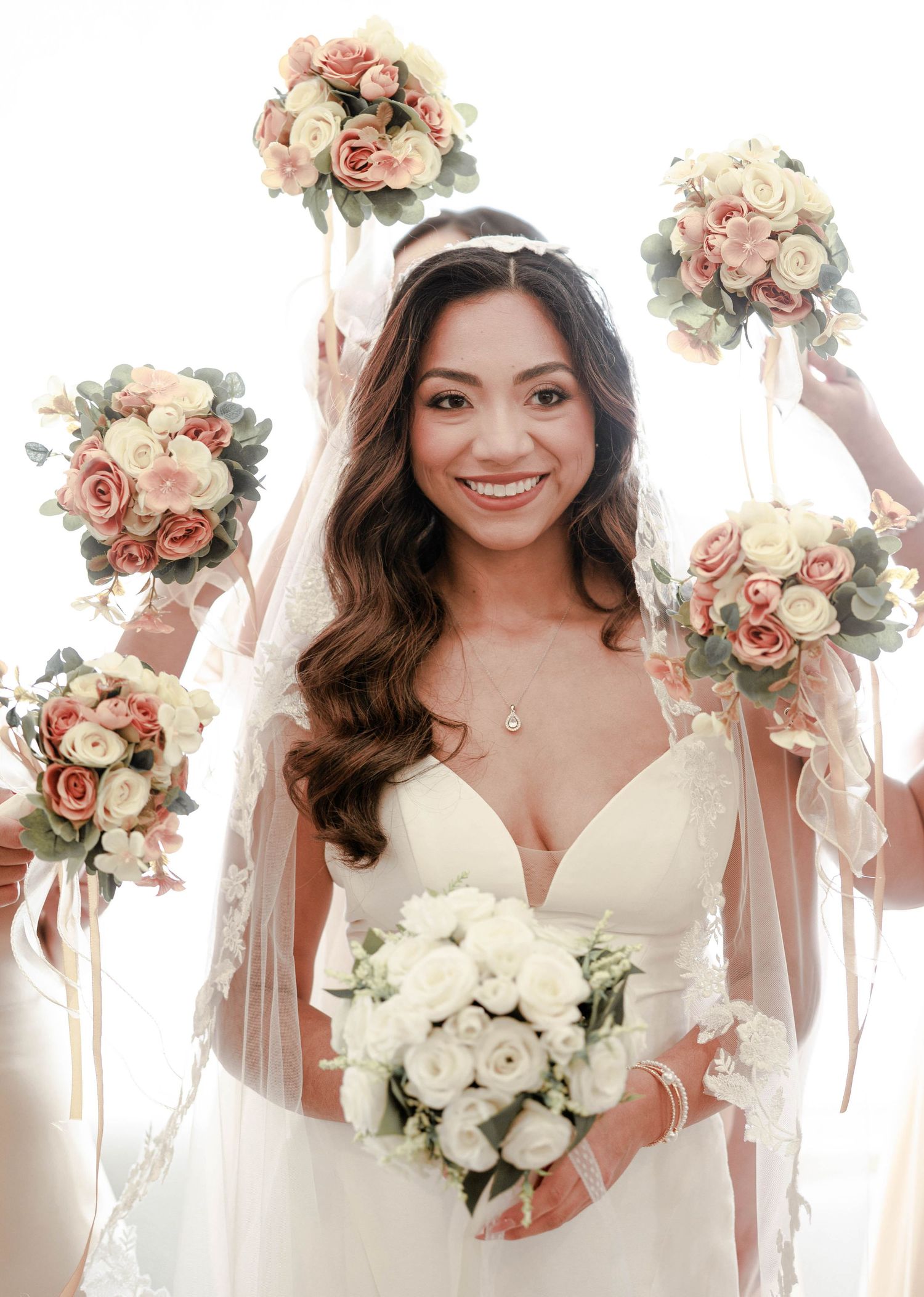 Smiling bride holds white bouquet surrounded by bridesmaids raising pink and cream rose bouquets.