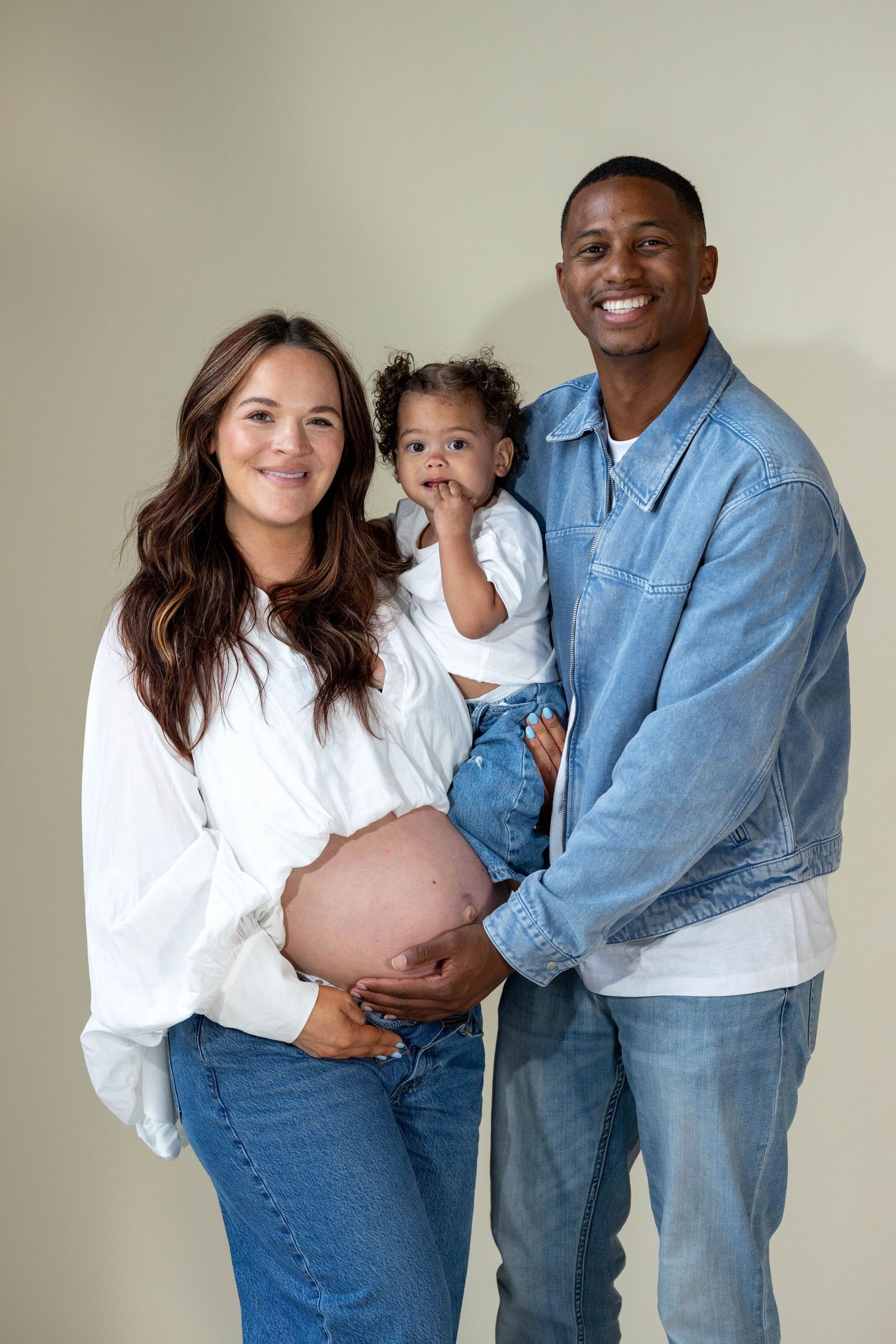 Smiling pregnant woman in white and partner in denim jacket hold their curly-haired toddler in studio.