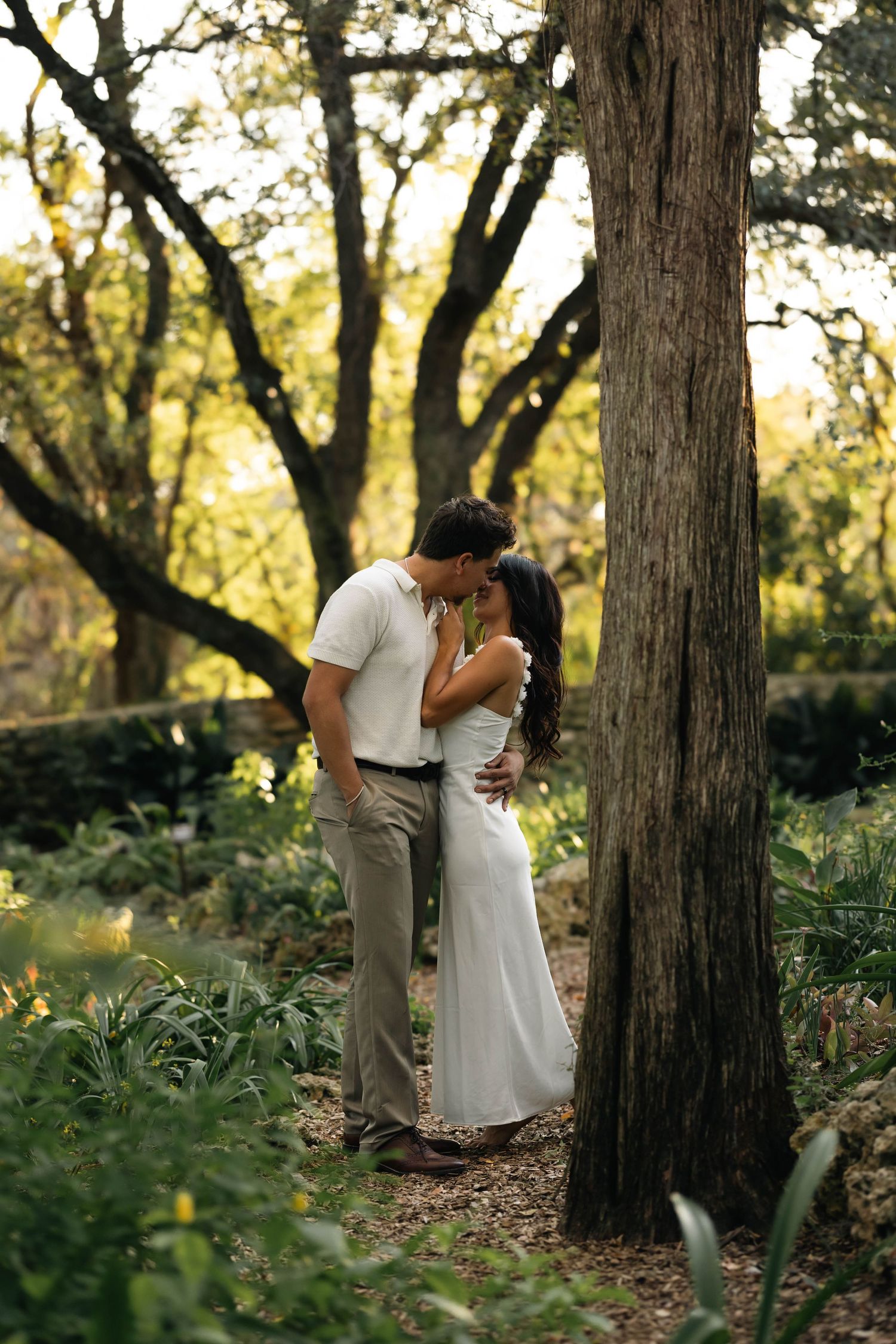 Couple sharing a romantic kiss near a large tree in a lush green garden.