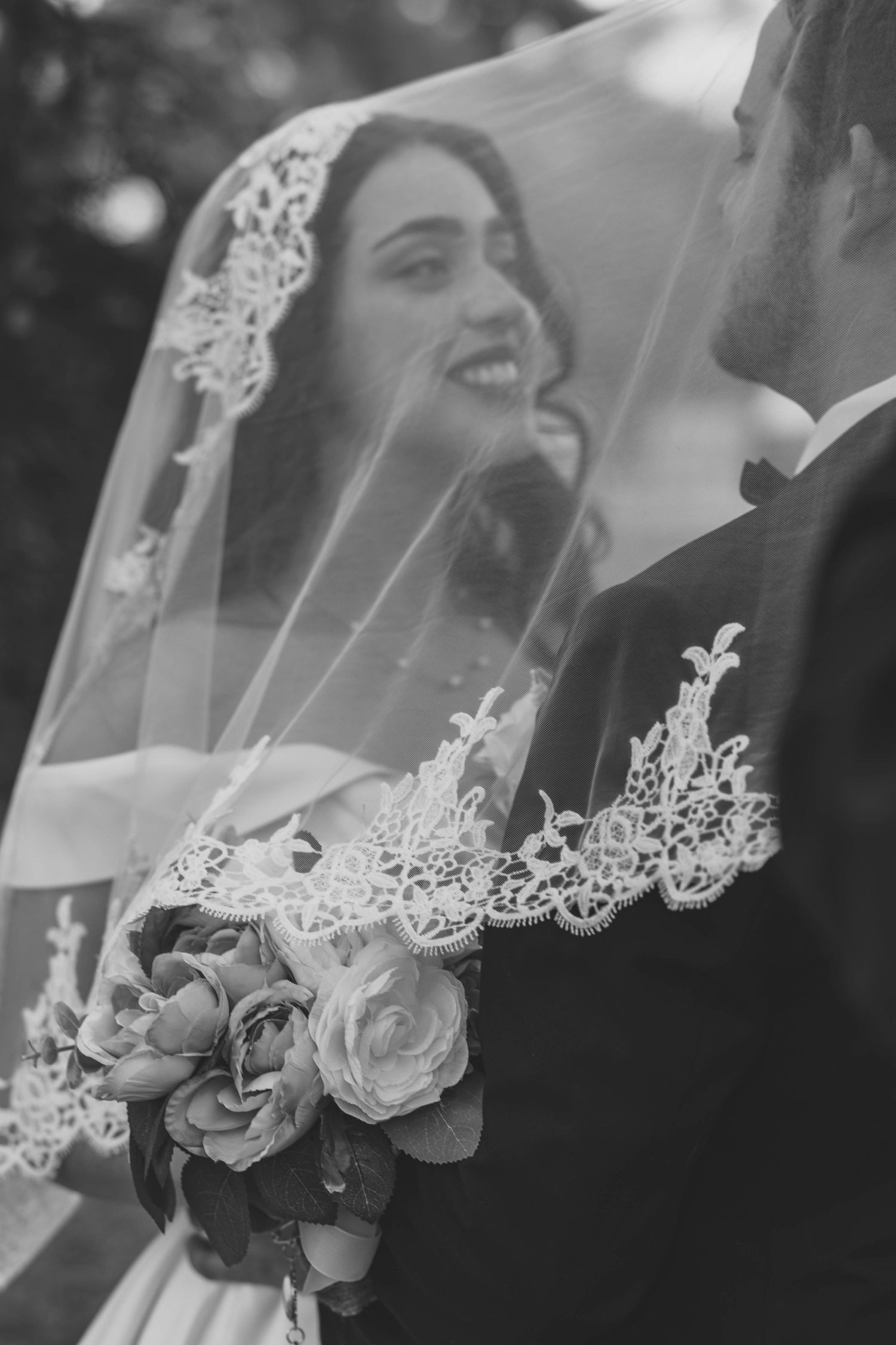 Black and white romantic portrait of bride and groom under lace veil with floral bouquet.