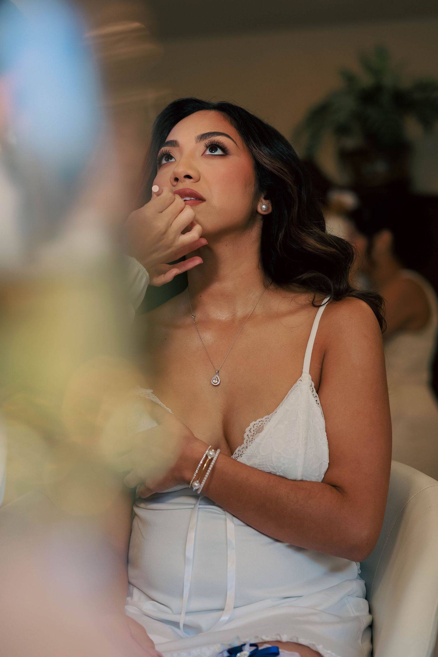 Elegant bride in white lace dress gazing upward thoughtfully, wearing pearl earrings and delicate necklace.