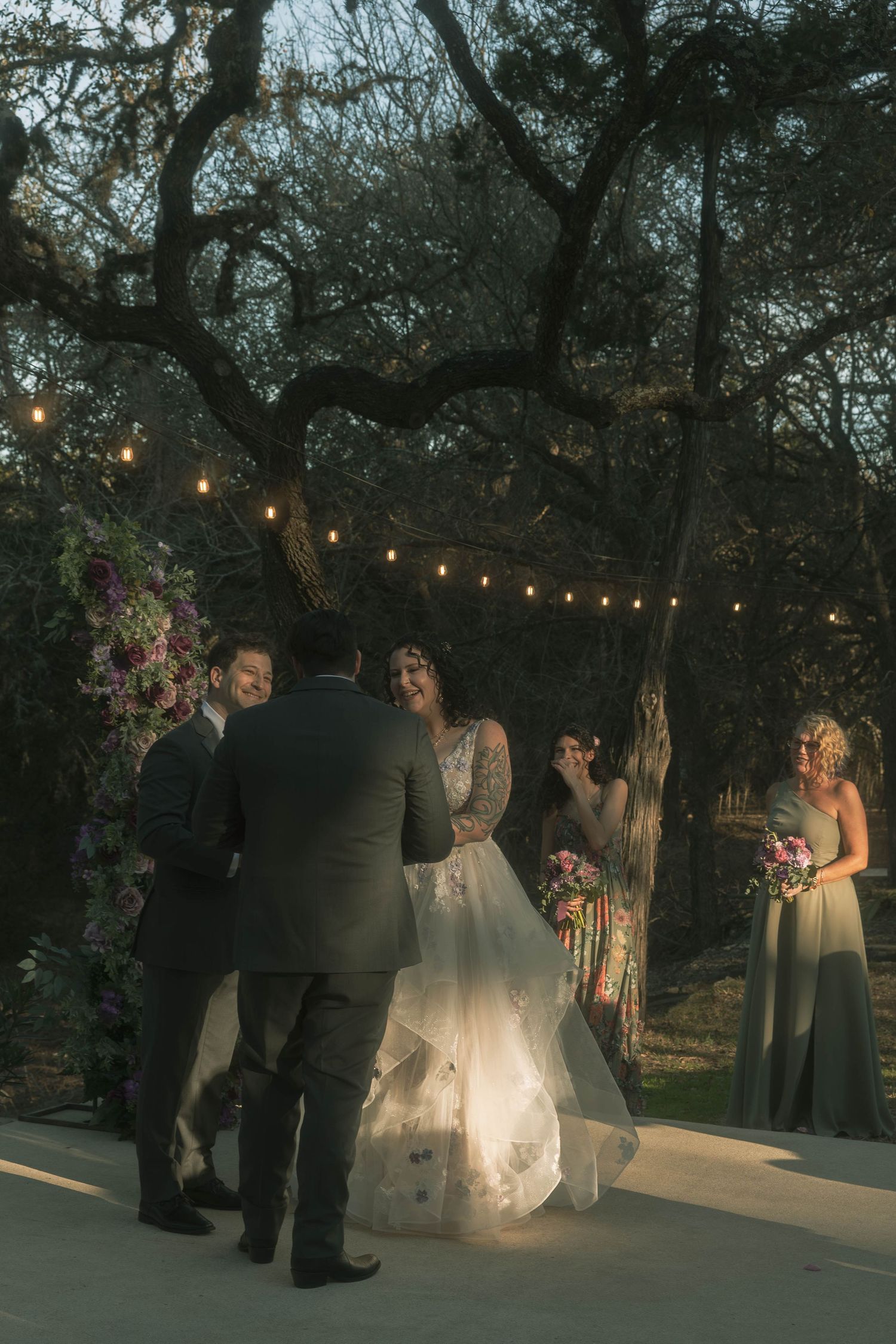 Bride and groom exchange vows outdoors at twilight under string lights and a floral arch surrounded by wedding party.