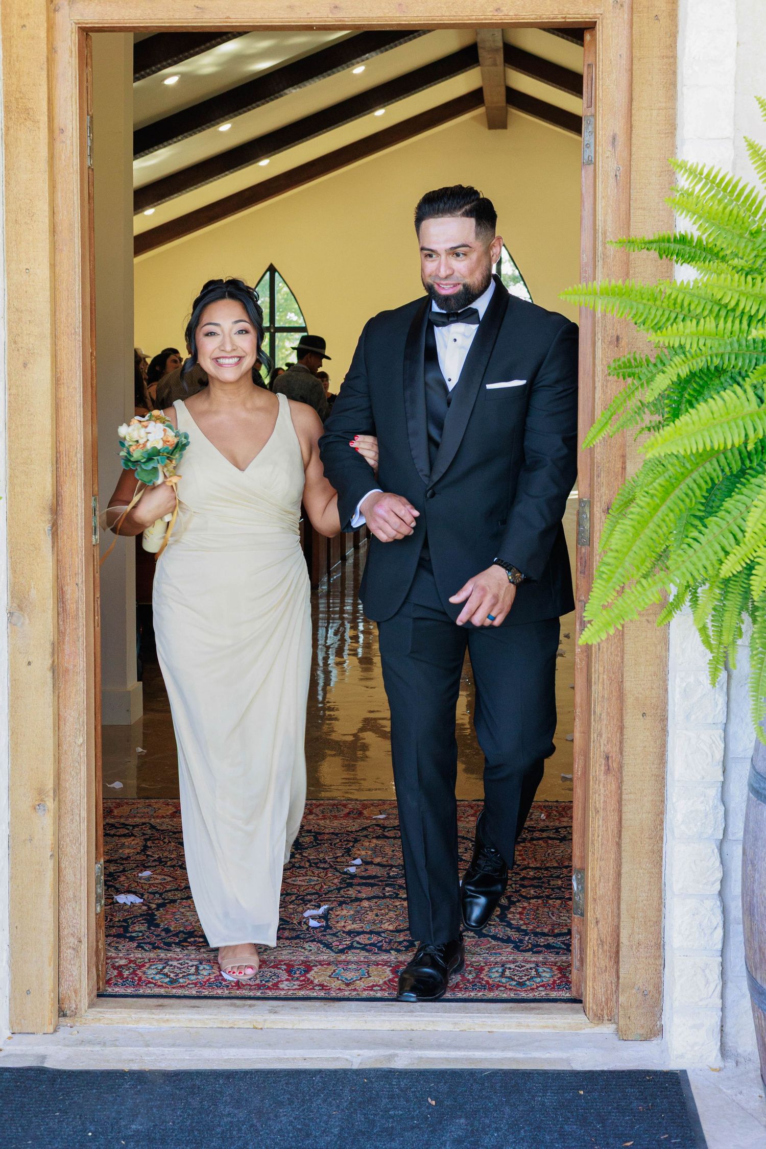 Smiling bridesmaid in champagne gown and groomsman in black tuxedo exit church ceremony doors.