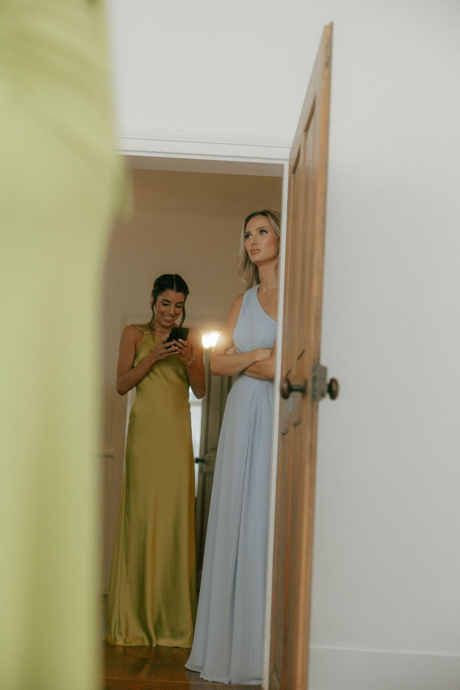 Two bridesmaids in green and blue satin gowns prepare behind a wooden door before the wedding.