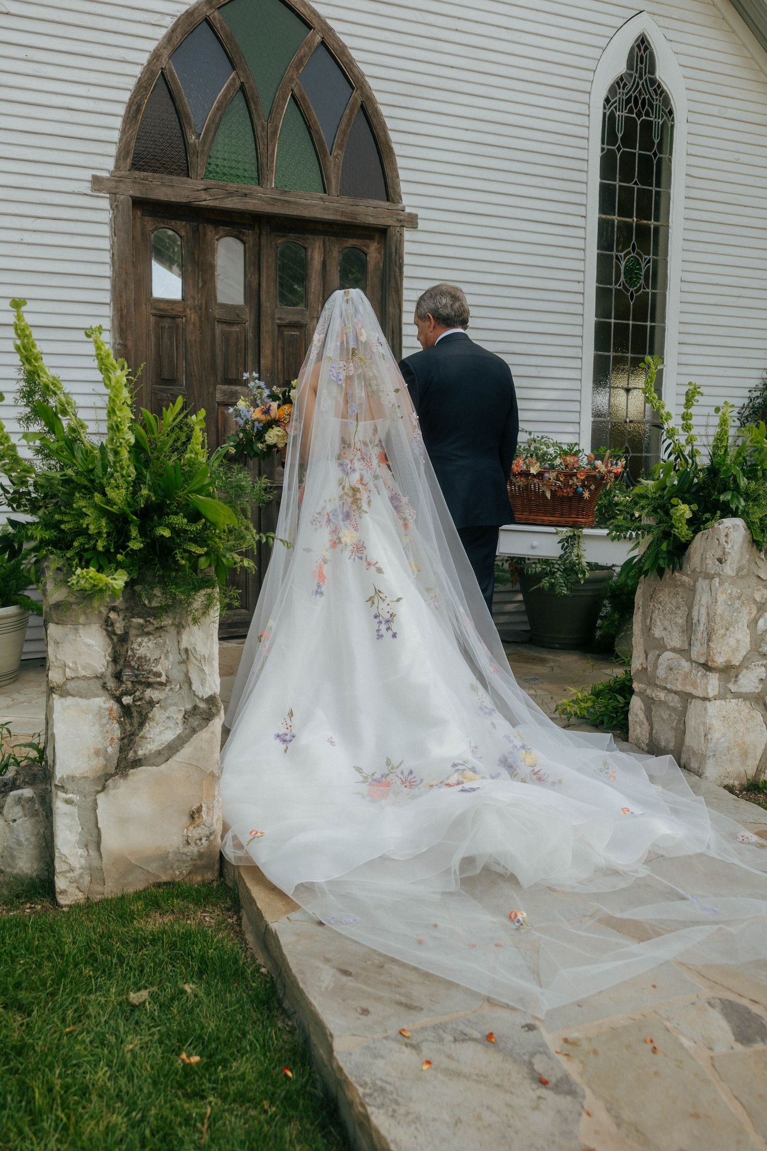 Bride in floral embroidered veil and gown stands outside a white chapel.