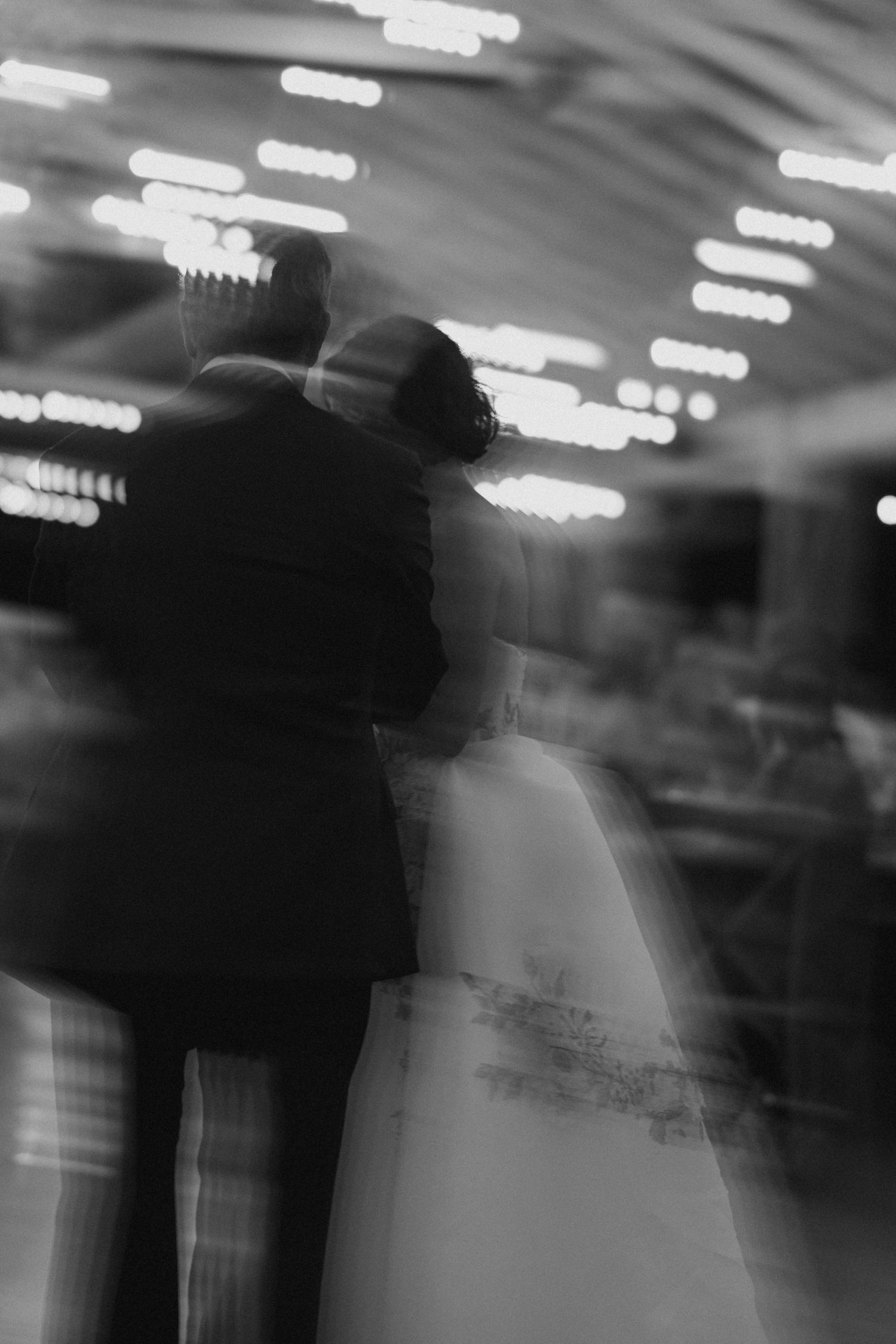 Artistic blurred black and white silhouette of bride and groom sharing their first dance under bright reception lights.