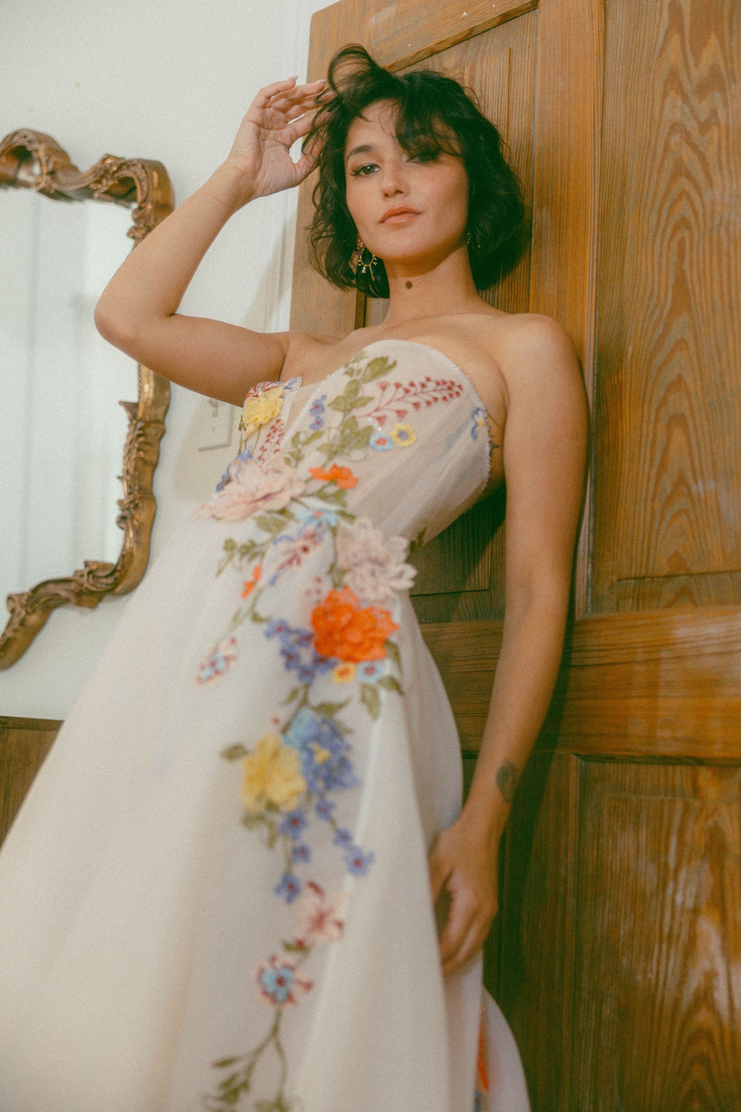 Woman in floral embroidered strapless white gown posing by wooden door with ornate mirror.