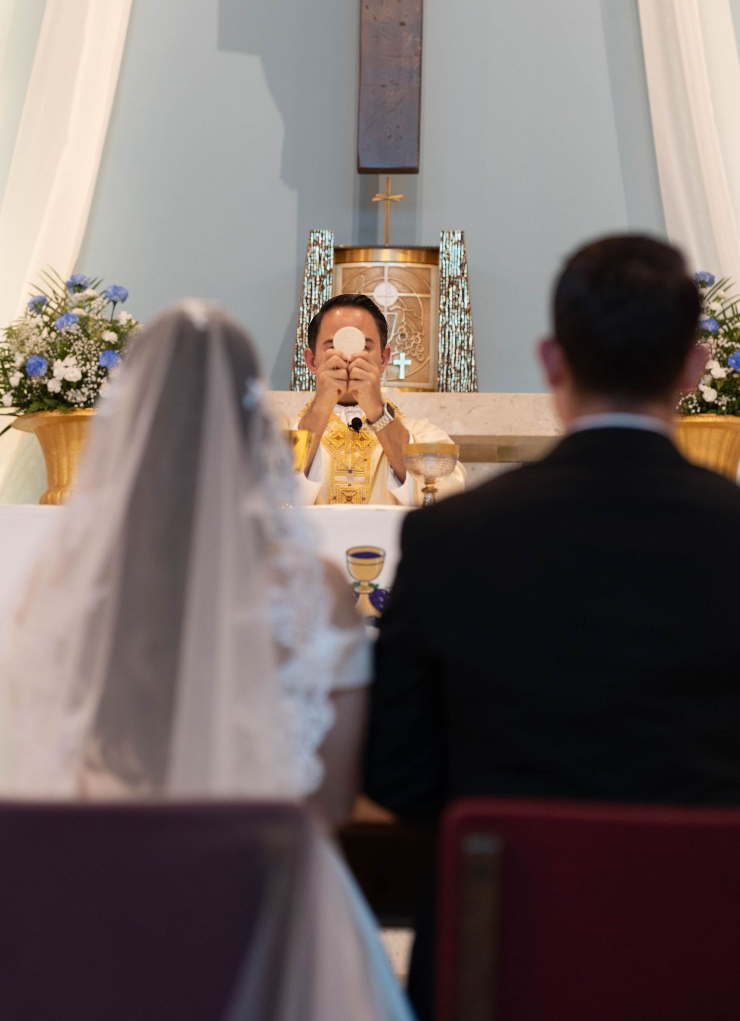 Priest elevates the Eucharist during a Catholic wedding ceremony as bride and groom kneel at altar.