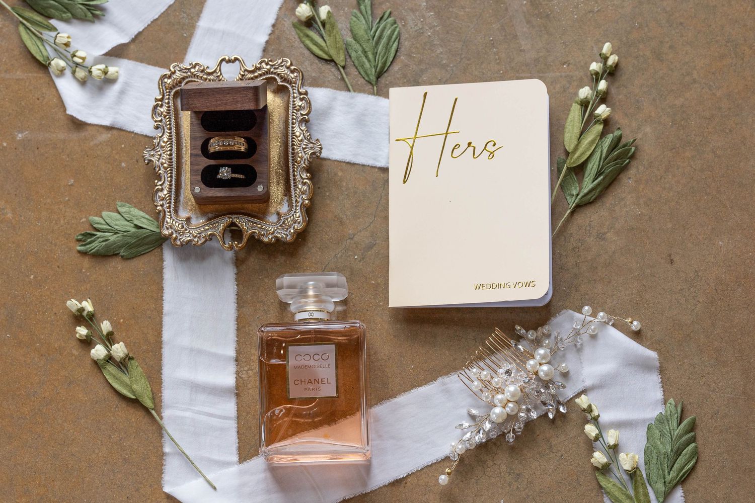 Wedding flat lay featuring rings in a vintage box, Chanel perfume, a Hers vow book, and floral accents.