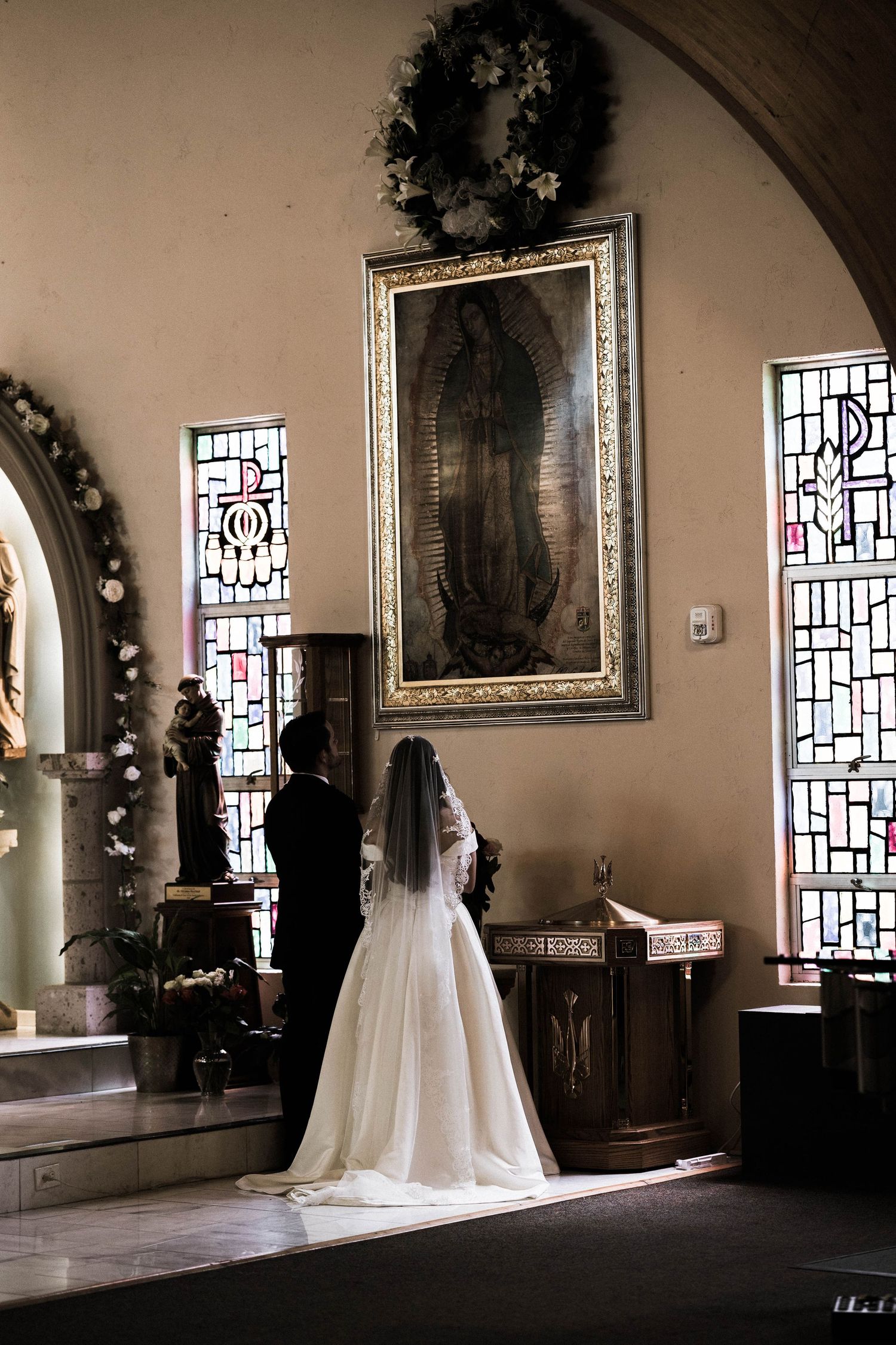 Bride and groom pray together before a Virgin of Guadalupe painting in a decorated Catholic church.