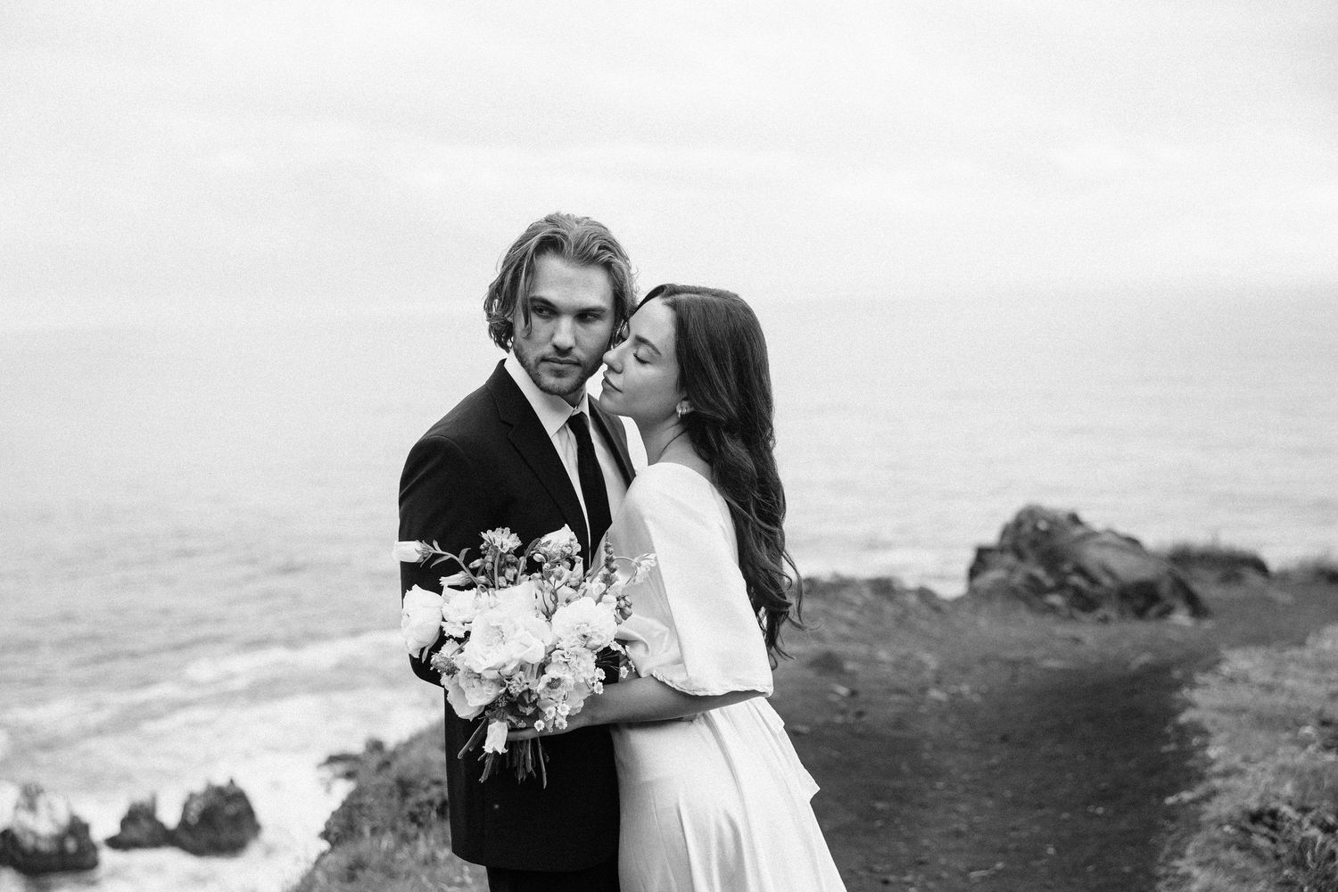 Black and white photo of bride tenderly kissing groom's cheek on a dramatic rocky oceanside cliff path.