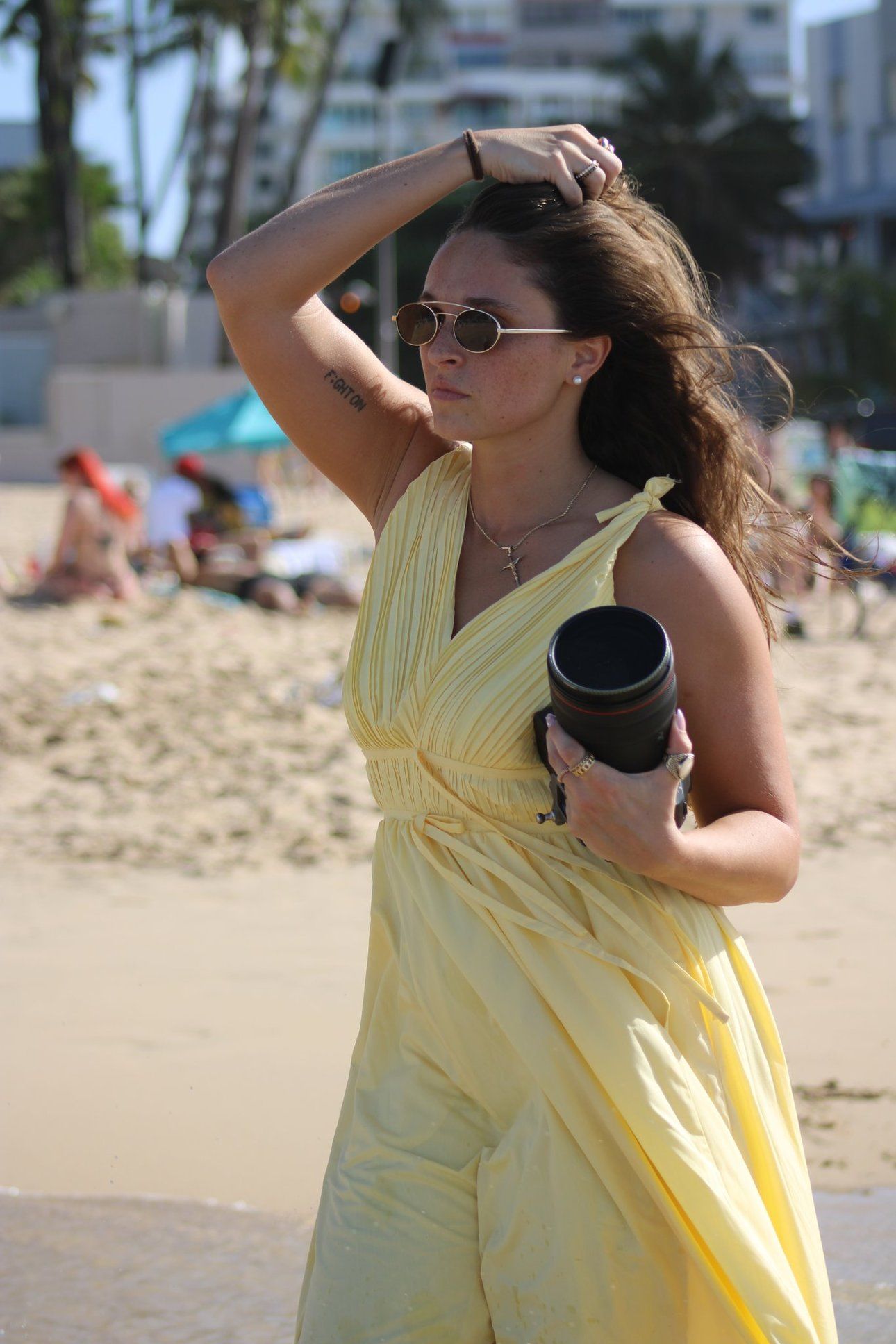 Female wedding photographer in a yellow dress holds a camera lens on a sunny beach.