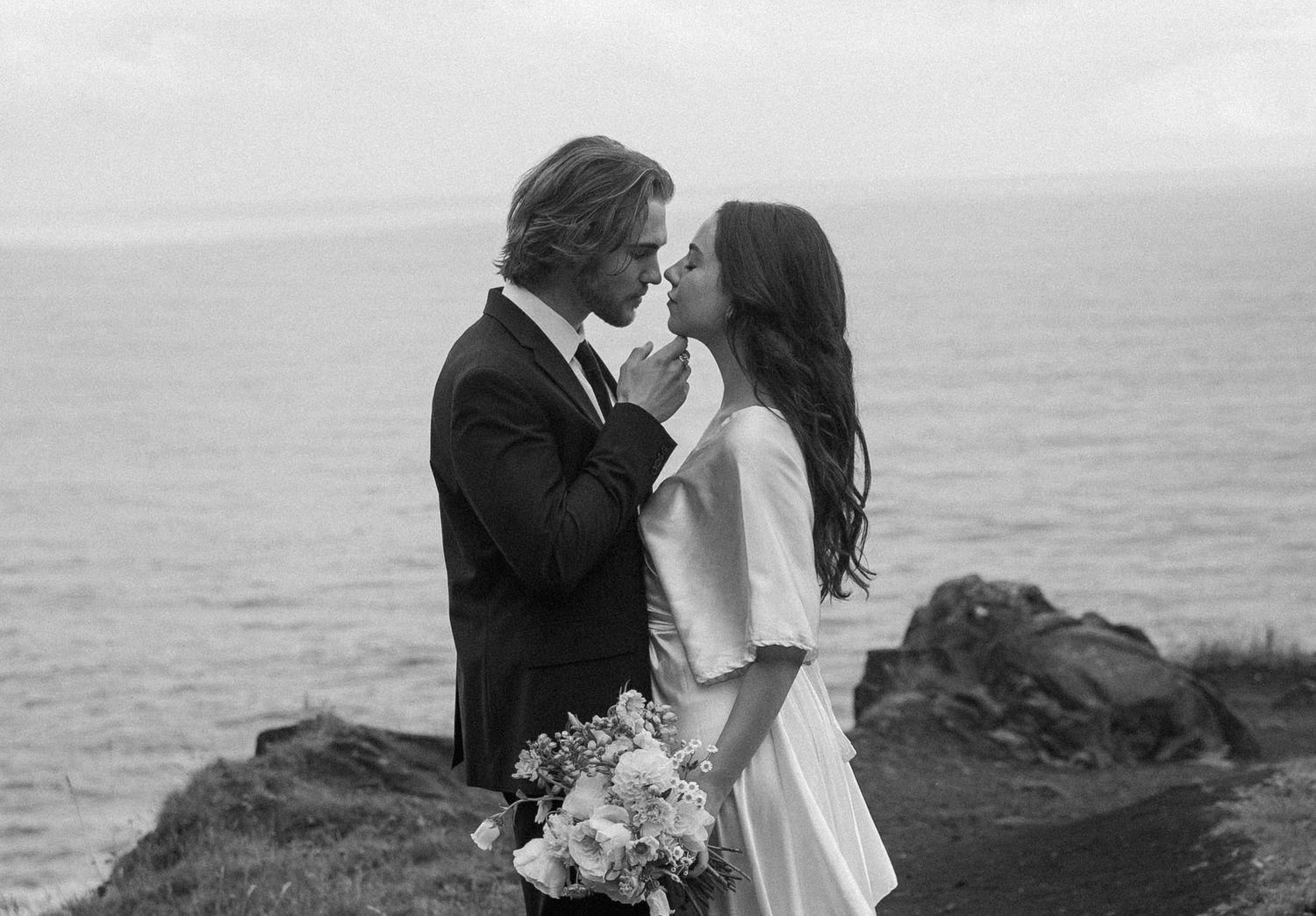 Bride and groom share a kiss on a dramatic coastal cliff in a black and white wedding photo.