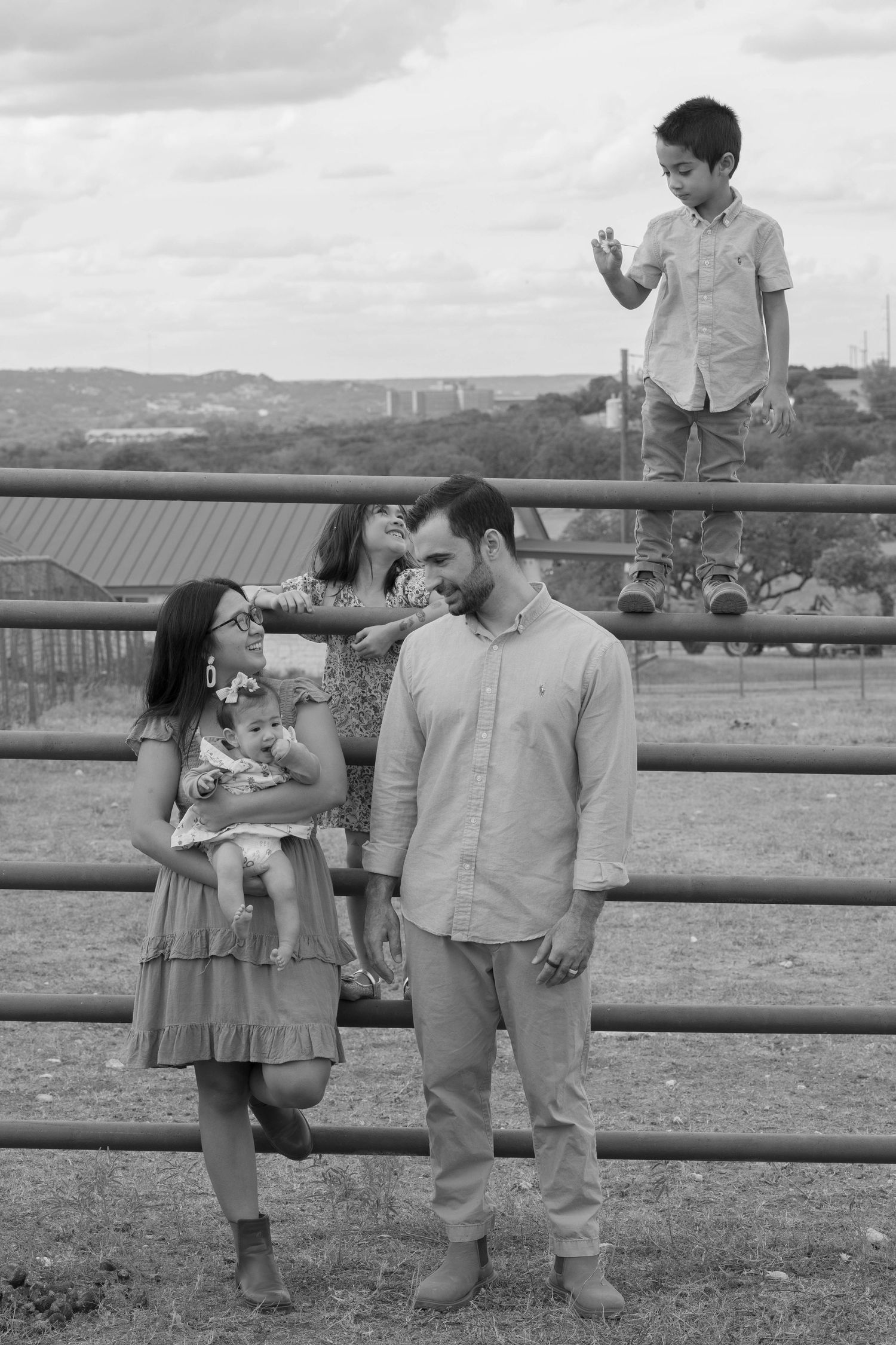 Black and white family portrait at a ranch fence with parents, two daughters, and son climbing the rails.