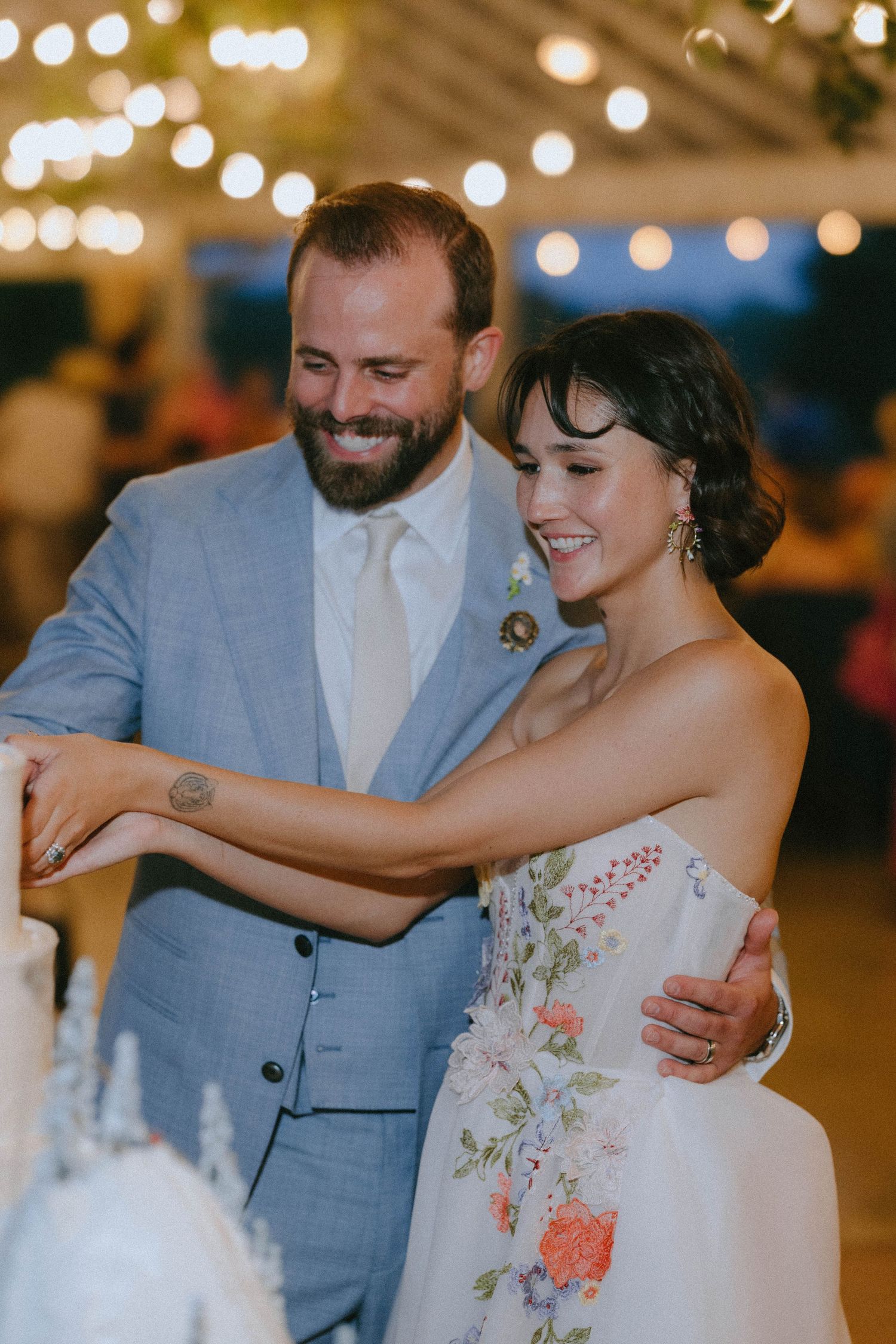Smiling bride and groom cutting their white wedding cake under string lights at reception.