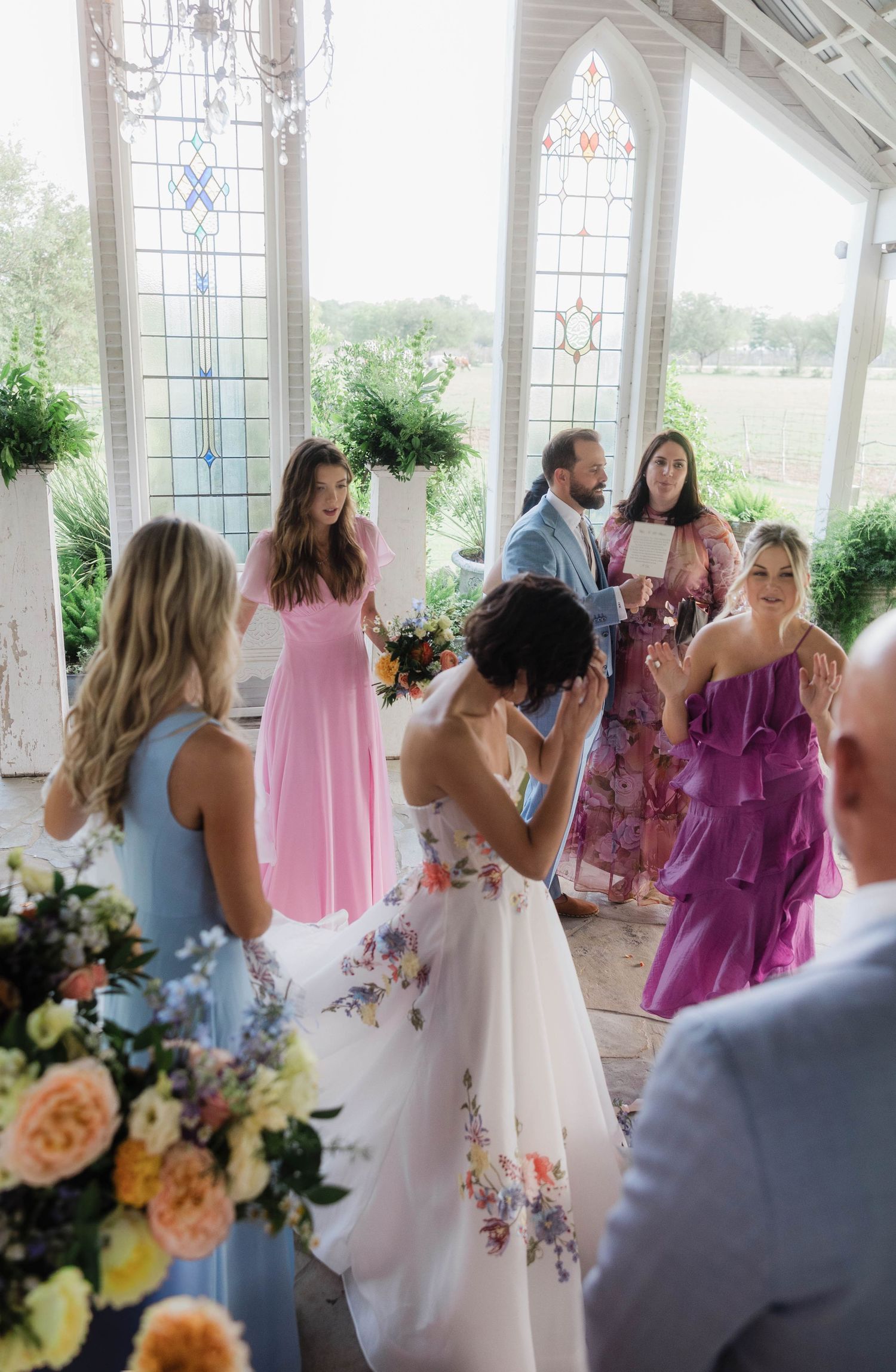 Bride in floral gown surrounded by bridesmaids in colorful dresses inside stained glass chapel.