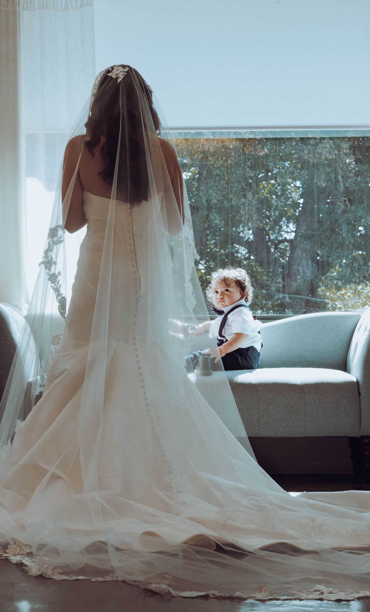 Bride in white gown with veil stands by window as toddler ring bearer sits on sofa.