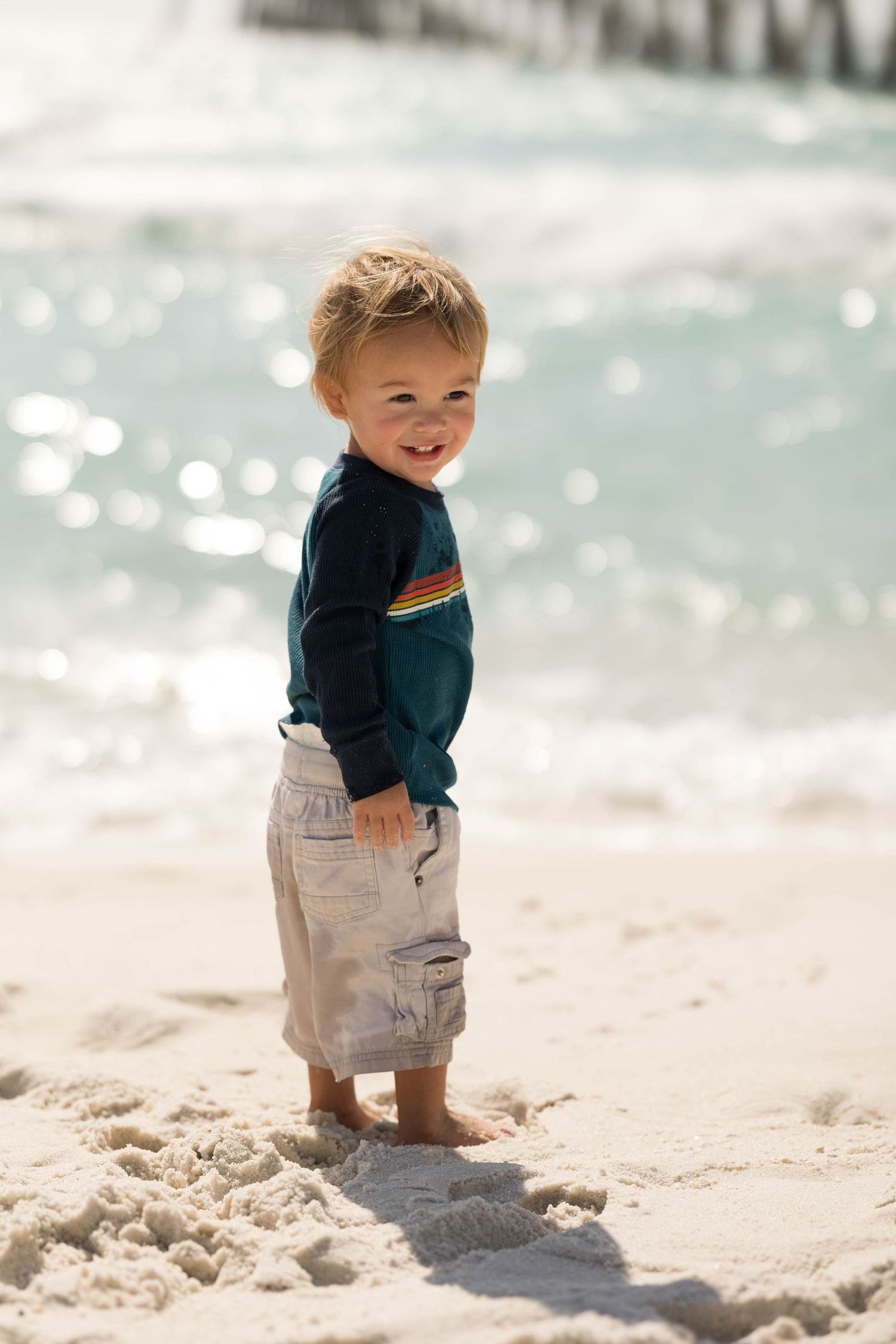 Smiling toddler boy standing barefoot on white sand beach wearing navy shirt and cargo shorts with ocean waves behind him.