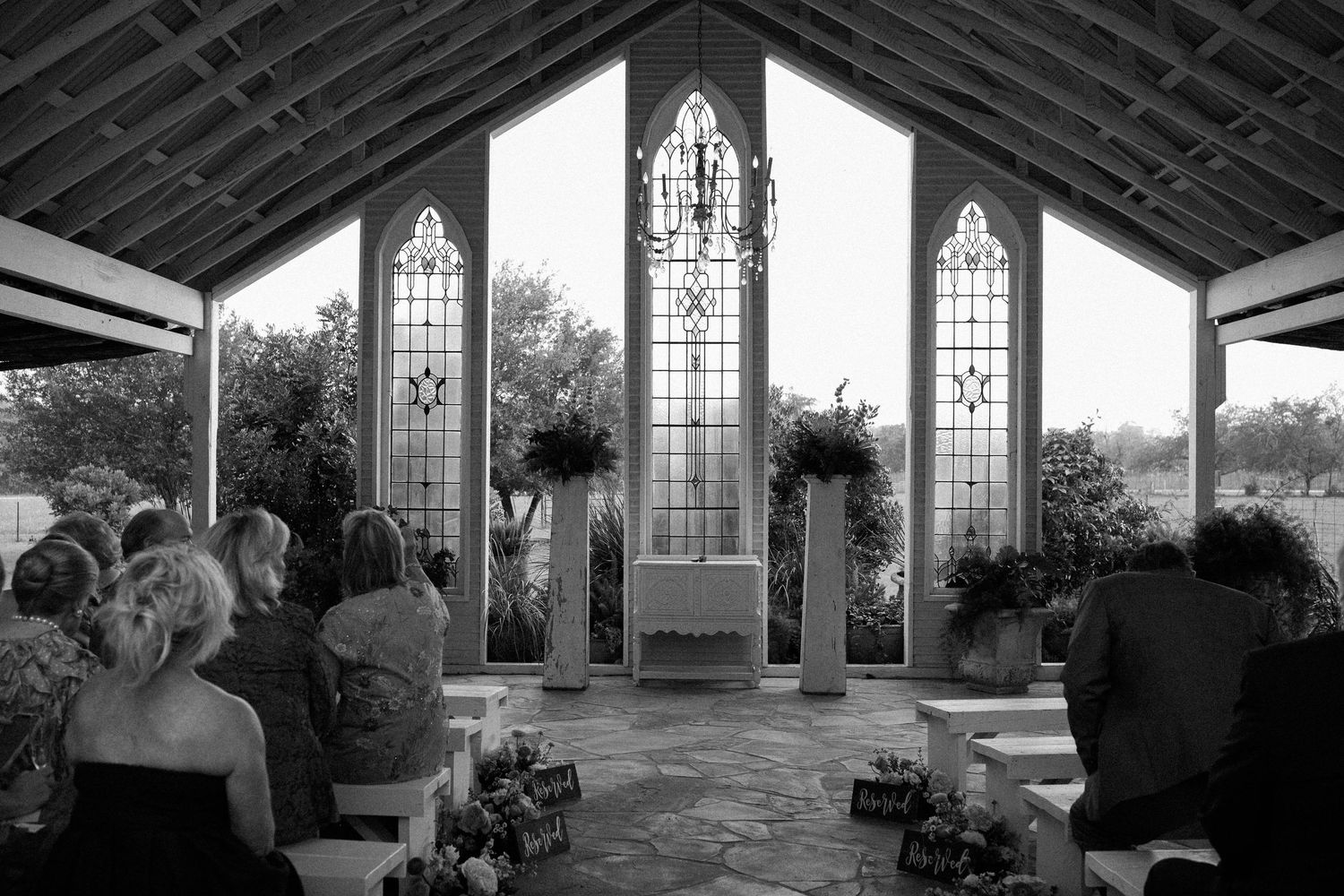 Black and white photo of a wedding ceremony inside a chapel with stained glass windows and guests seated in pews.