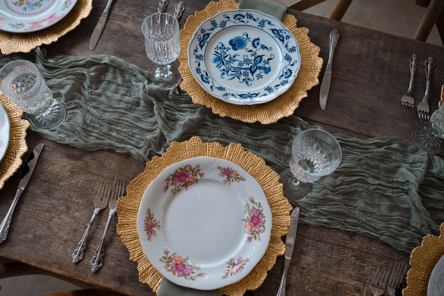 Overhead view of vintage mismatched china plates on gold chargers with sage green runner.